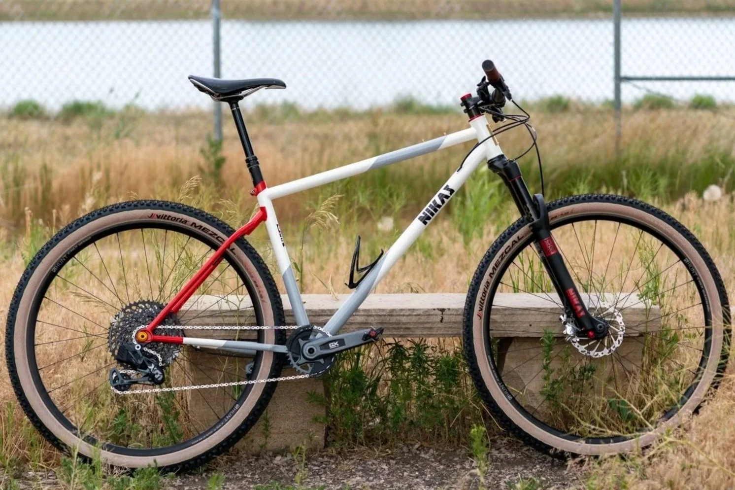 A modern mountain bike with a white and red frame, black handlebars, and thick tires, resting on a wooden bench in an outdoor grassy area.