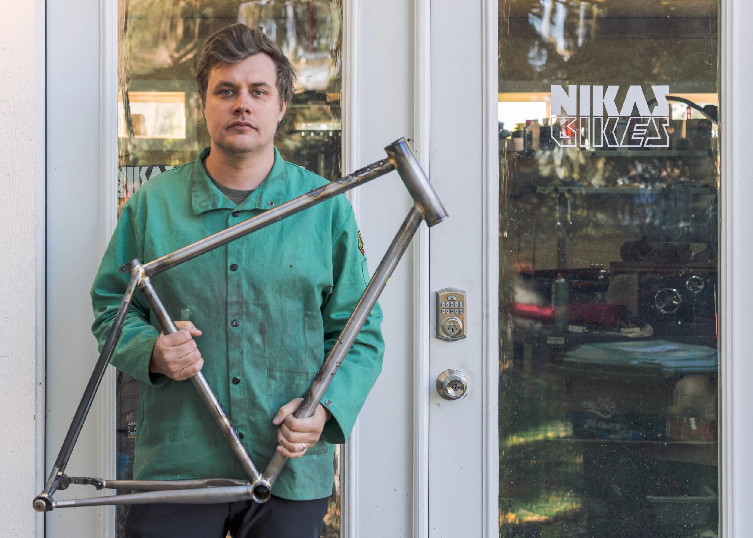 A man in a green jacket standing in front of a glass door holding a bicycle frame.