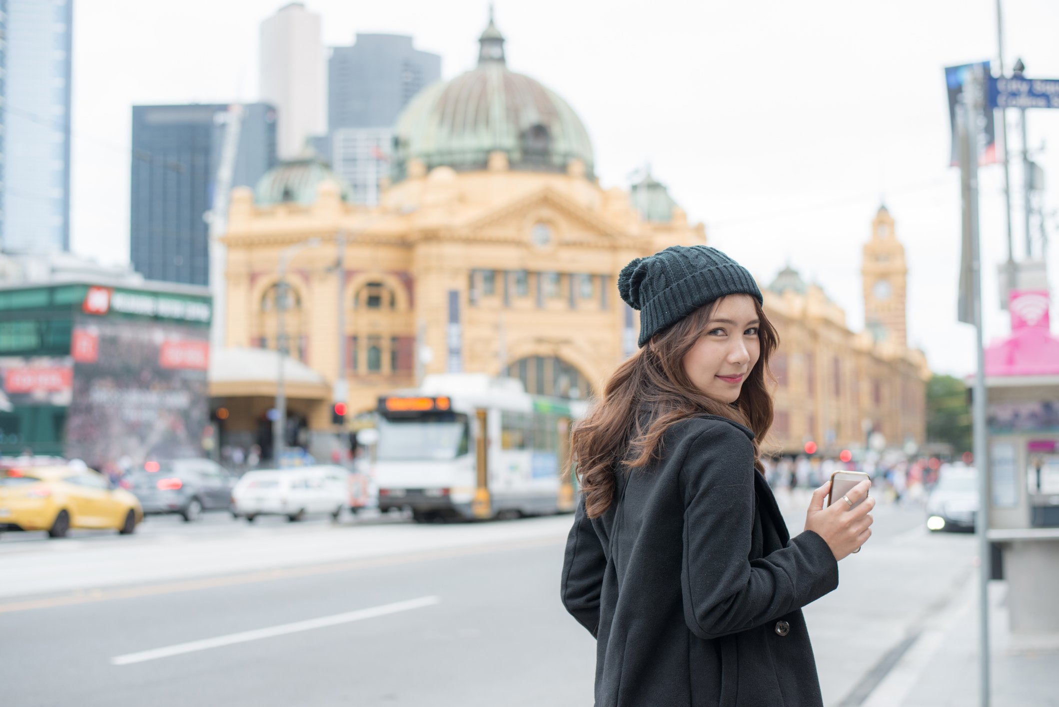 Portrait of an Asian woman traveling in Melbourne, standing outdoors with city landmarks in the background, highlighting the vibrant atmosphere of Victoria’s famously livable city.