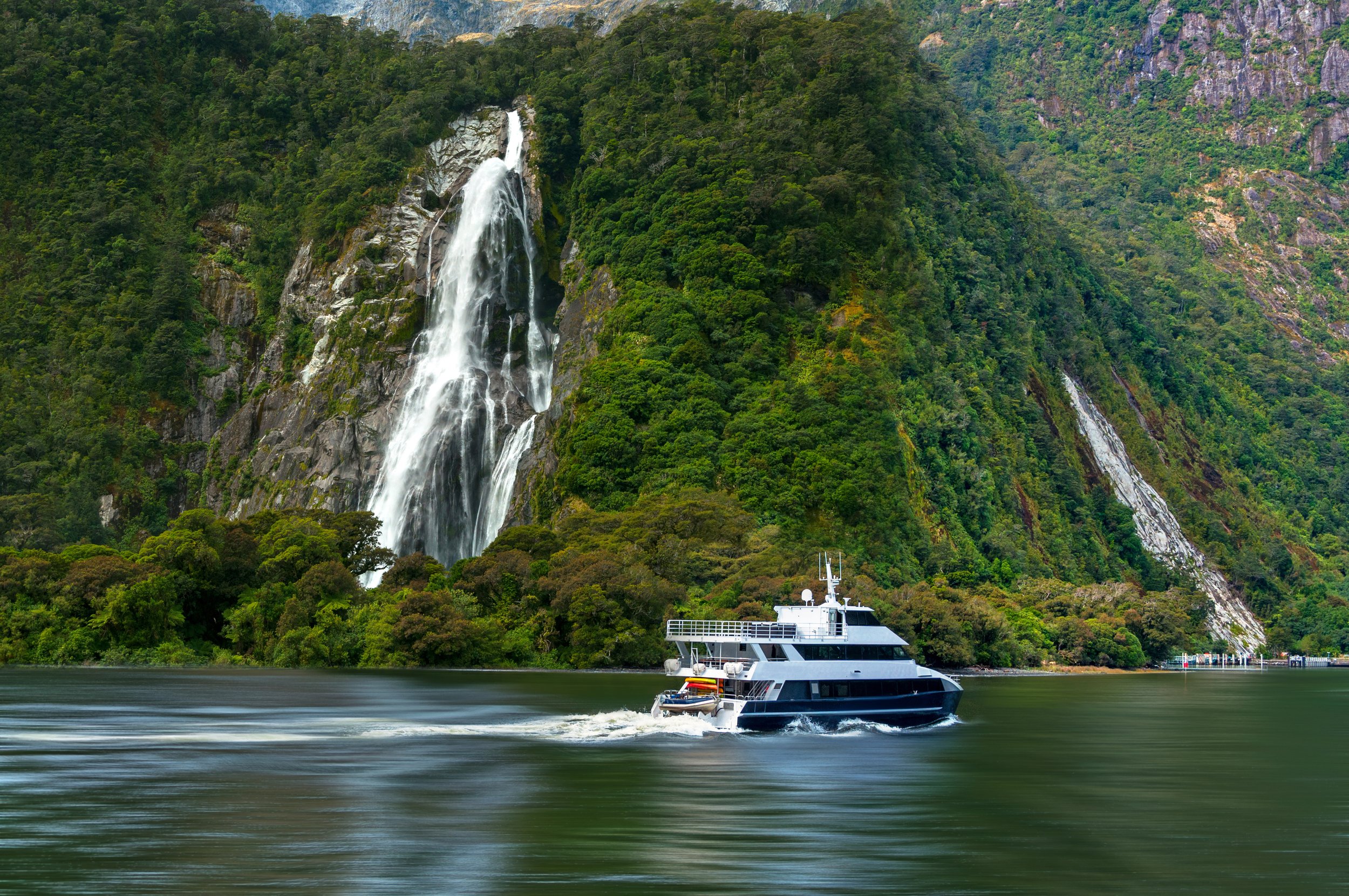 Luxury yacht cruising across a serene lake in Fiordland National Park, New Zealand, with a dramatic waterfall cascading down rugged mountain cliffs in the background.