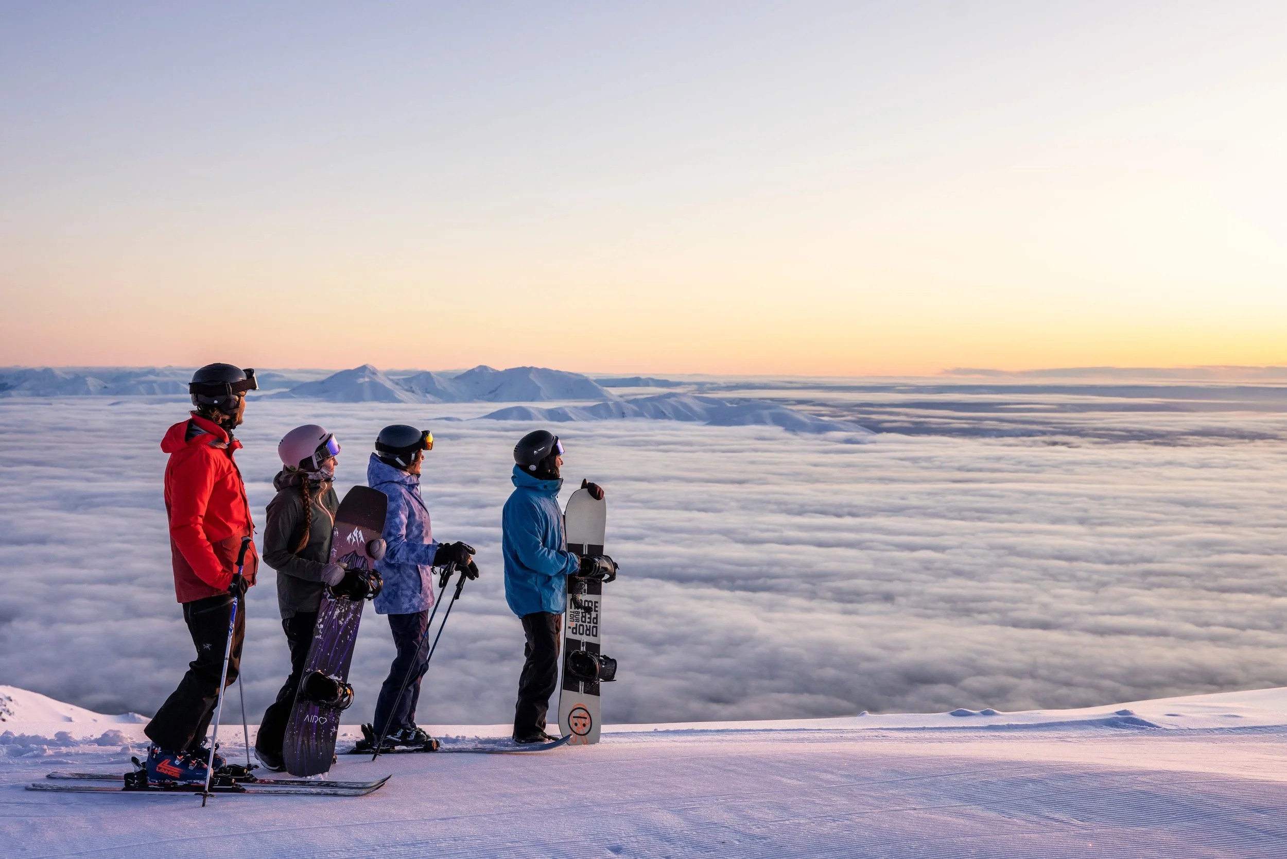 People standing on the snowy summit of Mount Hutt in Canterbury, silhouetted against a warm sunset with snow-covered peaks and sky glowing in the distance.