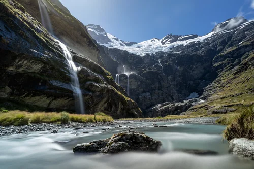 A long-exposure view of Earnslaw Burn Basin in New Zealand, showing silky blue-green glacial water flowing past rocks, waterfalls cascading down dark cliffs, and snow-covered peaks beneath a clear sky.