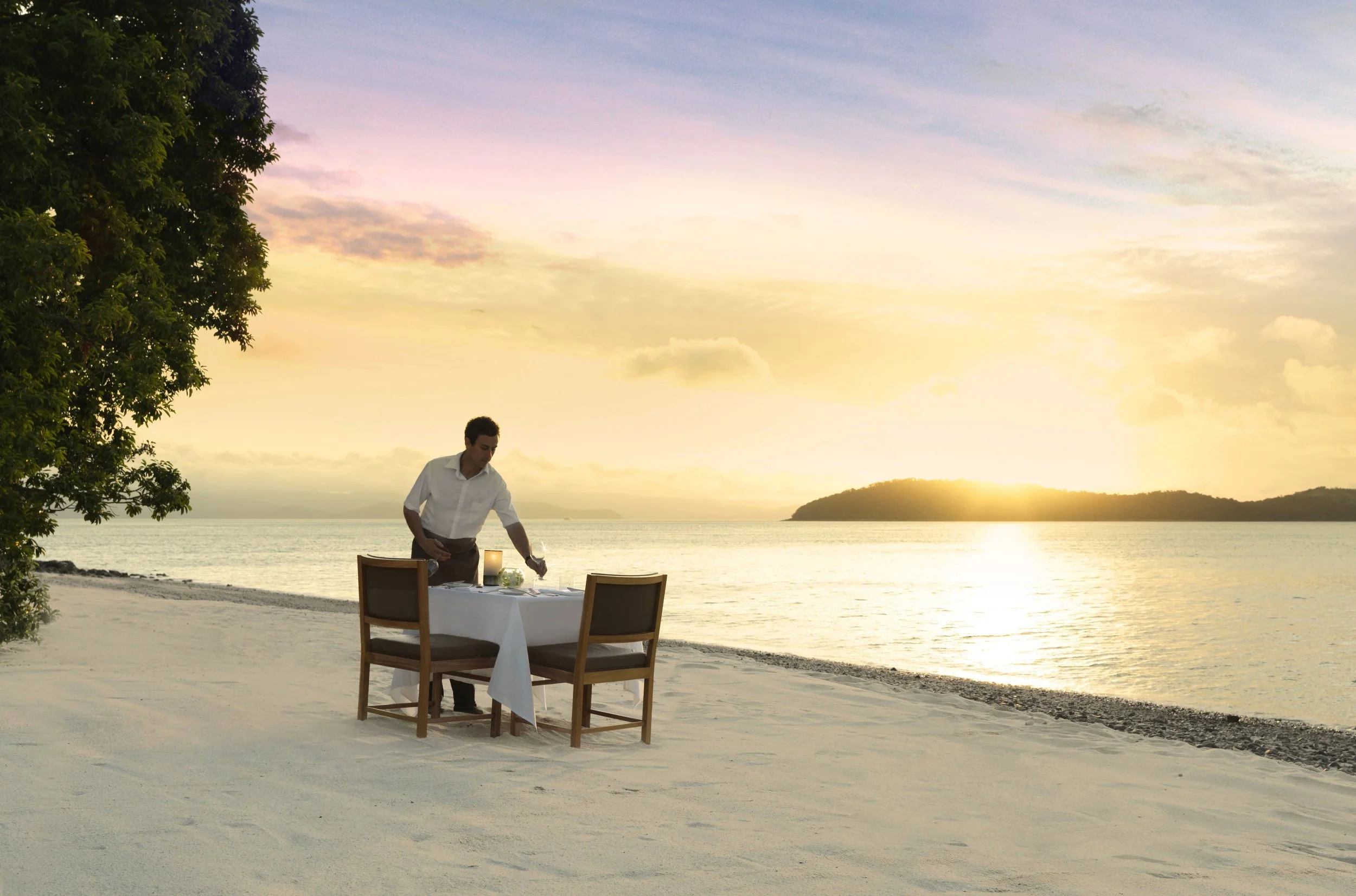 A private fine-dining experience set on Pebble Beach at Qualia resort, with an elegant table for two overlooking the water at sunset, offering gourmet cuisine against the backdrop of the Great Barrier Reef.