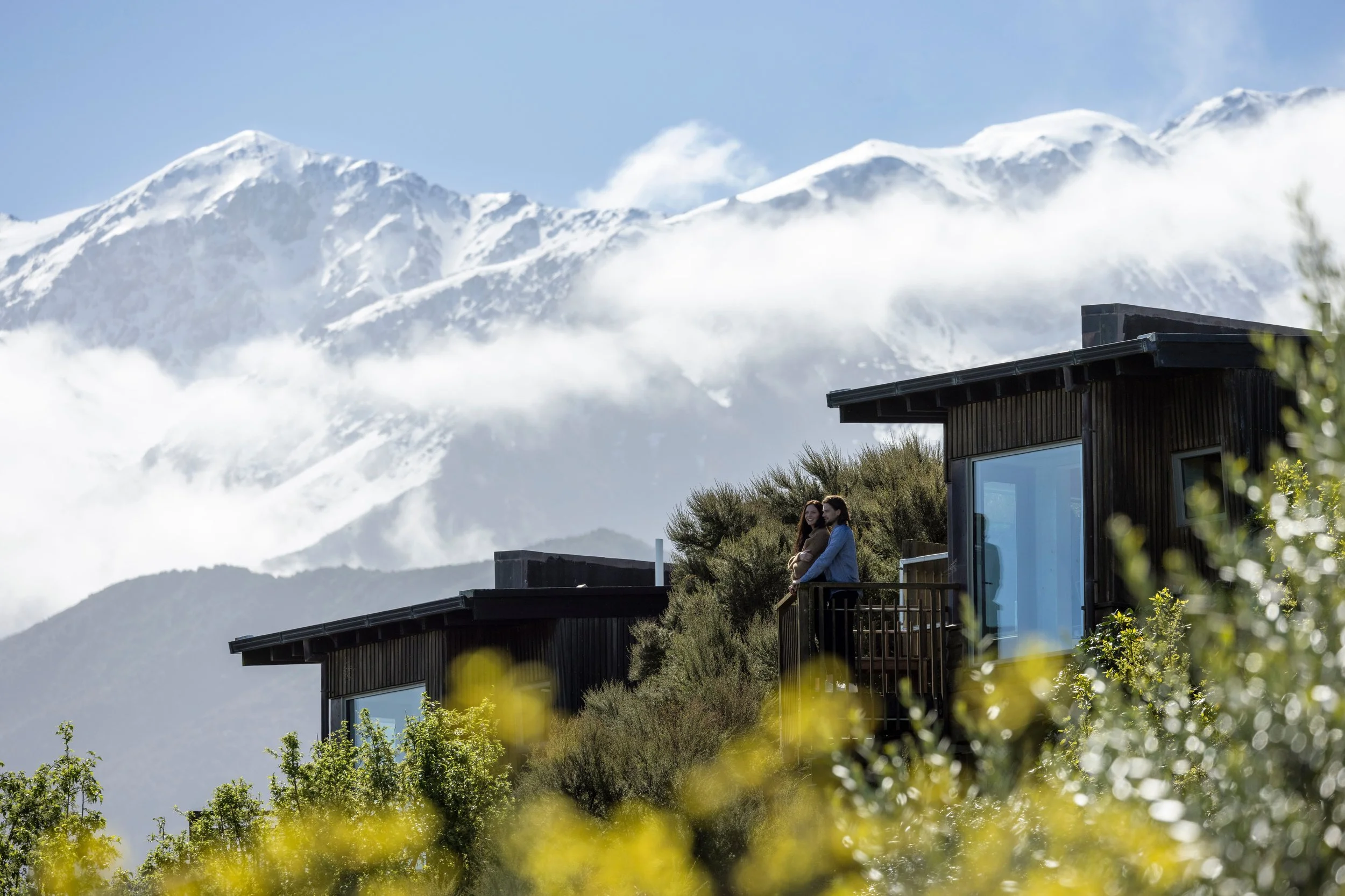 A couple enjoying the view from their luxury lodge in Kaikōura, Canterbury, with lush greenery in the foreground and snow-capped mountains in the background.