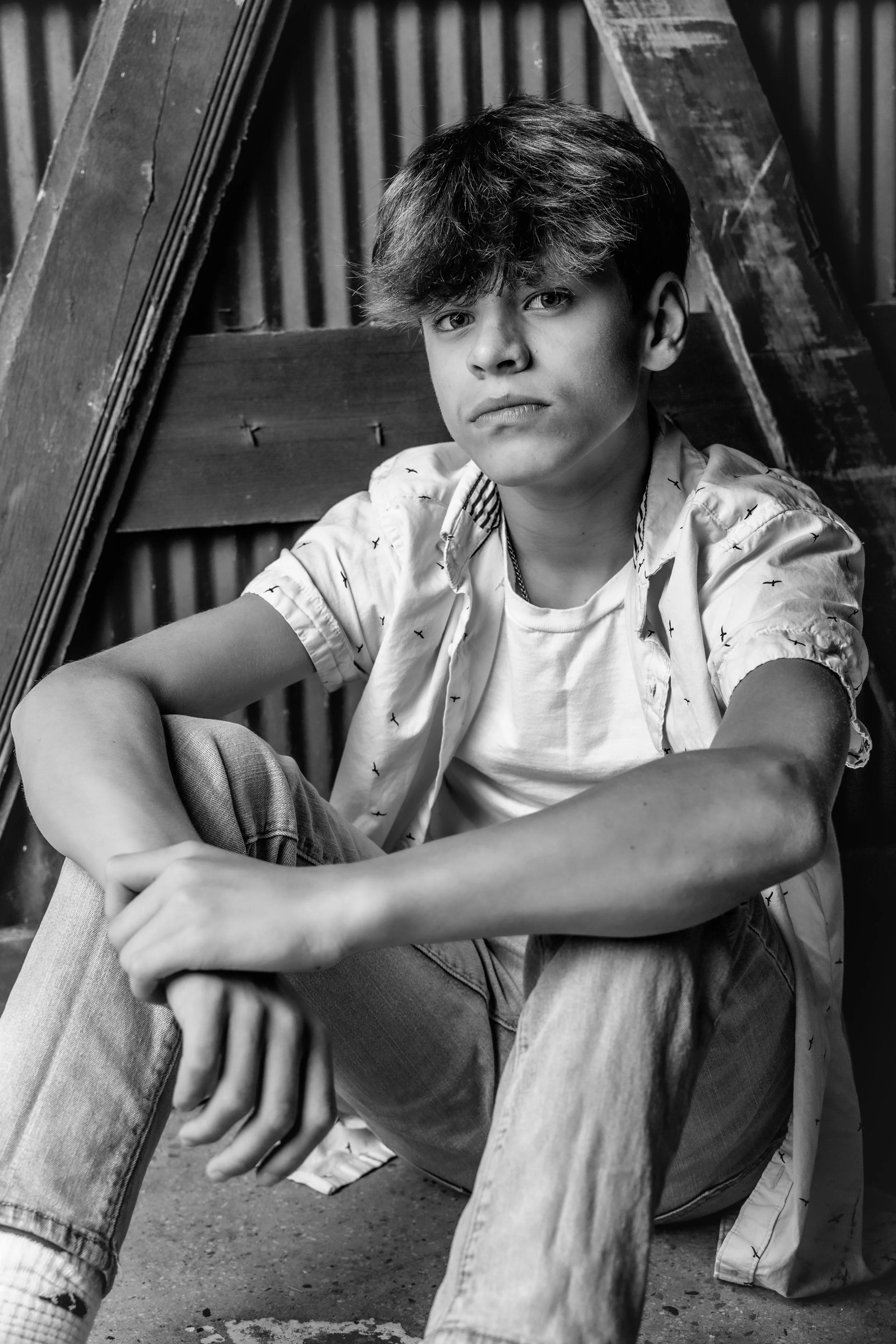 Young man sitting on the ground in front of a wooden backdrop with corrugated metal, wearing a patterned short-sleeve shirt and jeans, looking at the camera in black and white.