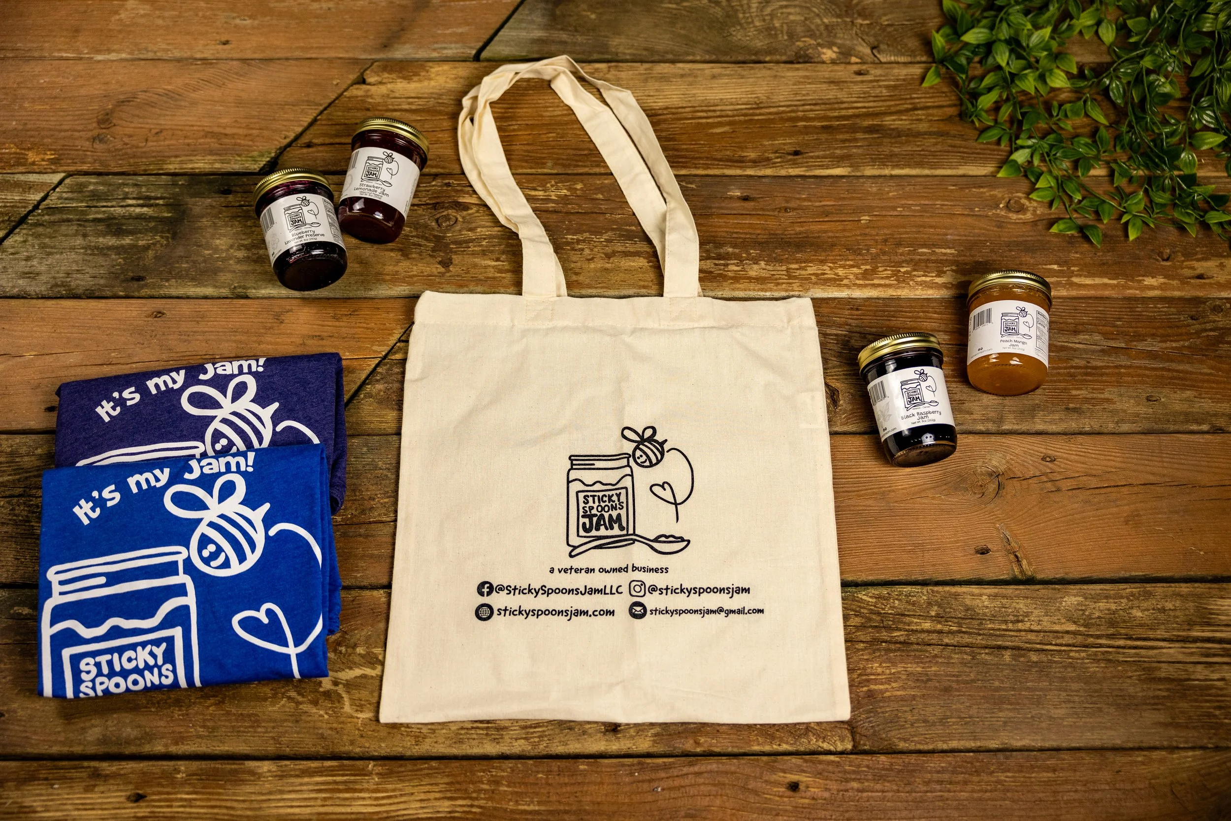A collection of jars of jam, a beige tote bag with branding, and folded blue and purple towels with jelly jar and bee illustrations on a wooden surface. Green foliage in the top right corner.