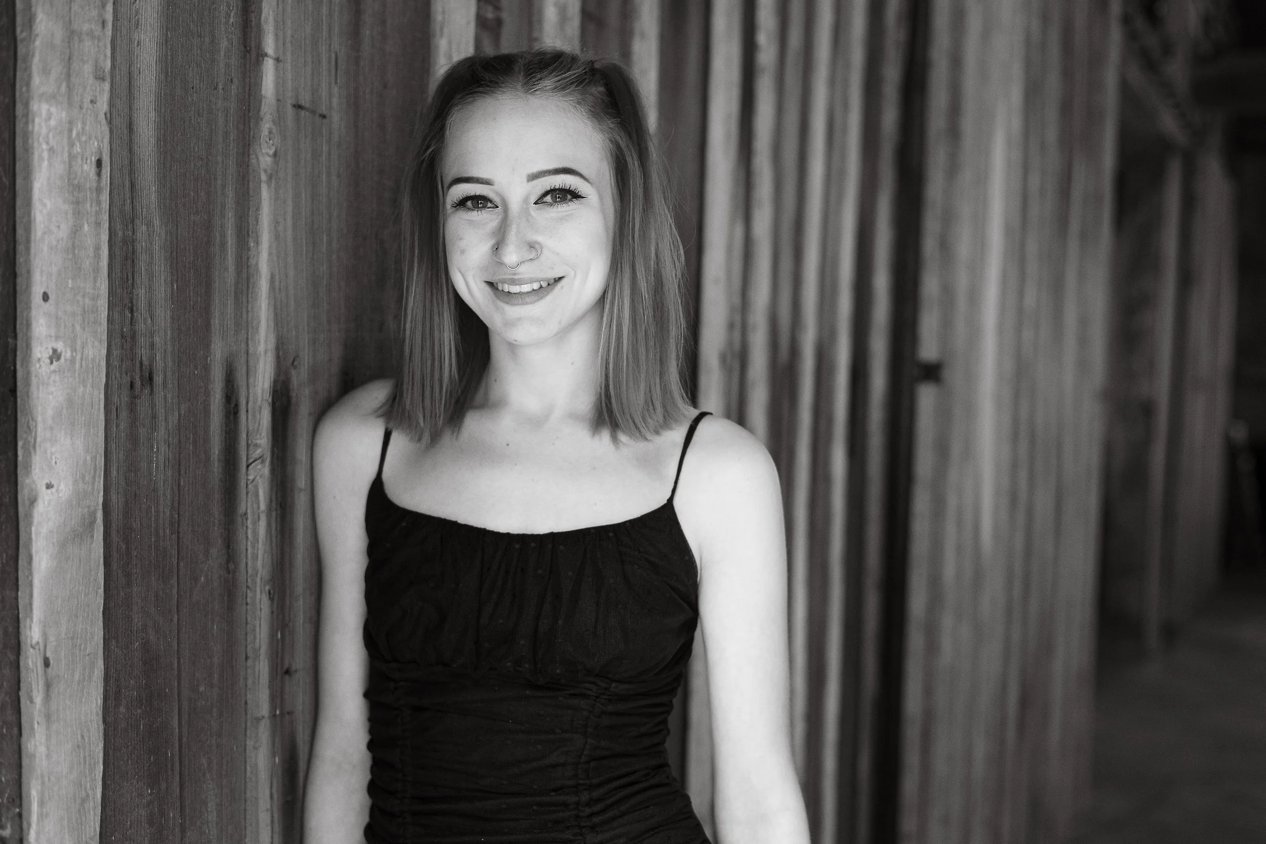 A young woman with shoulder-length hair, wearing a black spaghetti strap top, smiling, and standing against a wooden wall in black and white.
