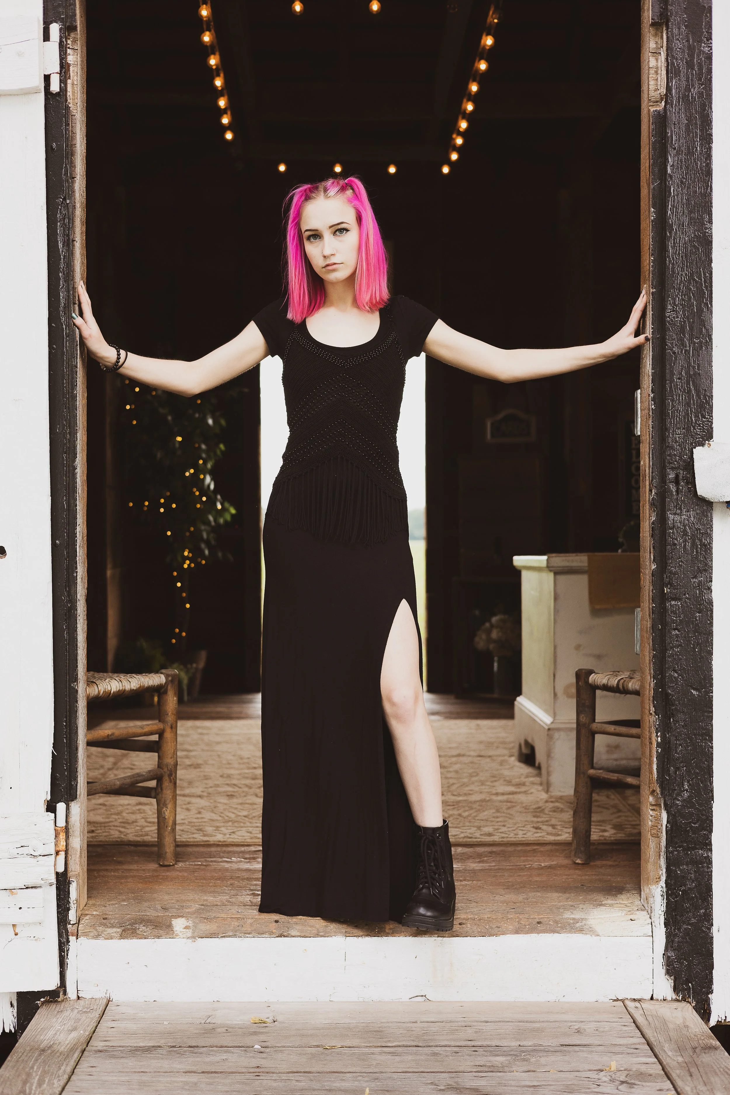 A young woman with pink hair wearing a black dress with a high slit stands in a doorway, holding onto the door frame, with string lights hanging from the ceiling behind her.