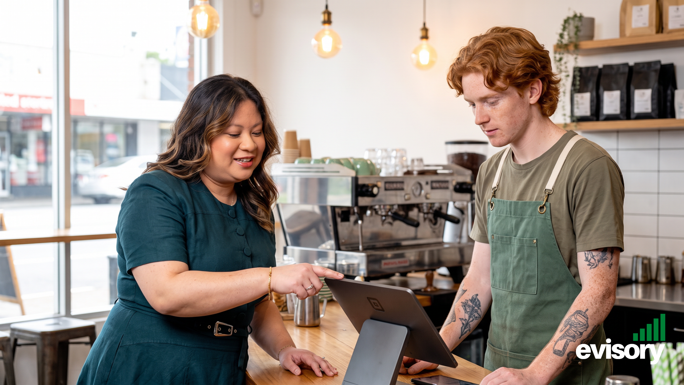 An Evisory hospitality accountant pointing at a tablet POS terminal on a café counter while assisting a barista in reviewing POS settings.