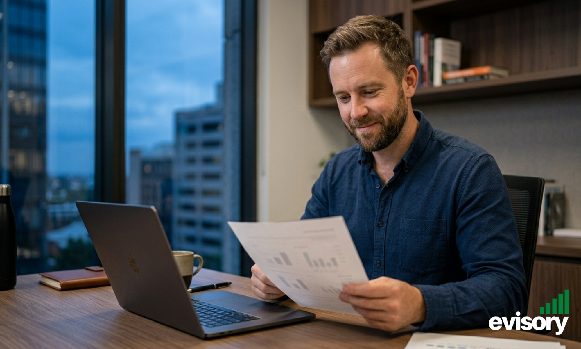 Australian man in his late 30s reviewing printed financial documents at a clean office desk with an open laptop beside him, appearing relieved and focused in natural window light.