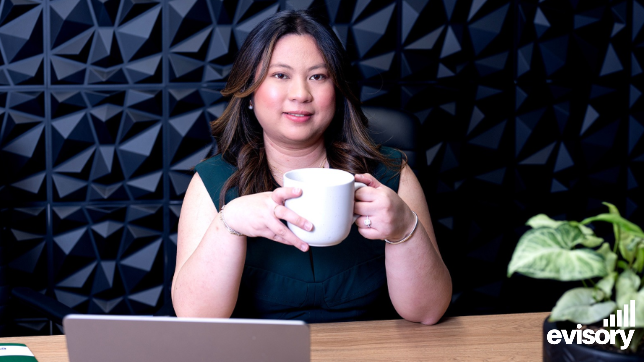 Professional woman holding a white coffee mug at an office desk with a laptop and the Evisory logo.