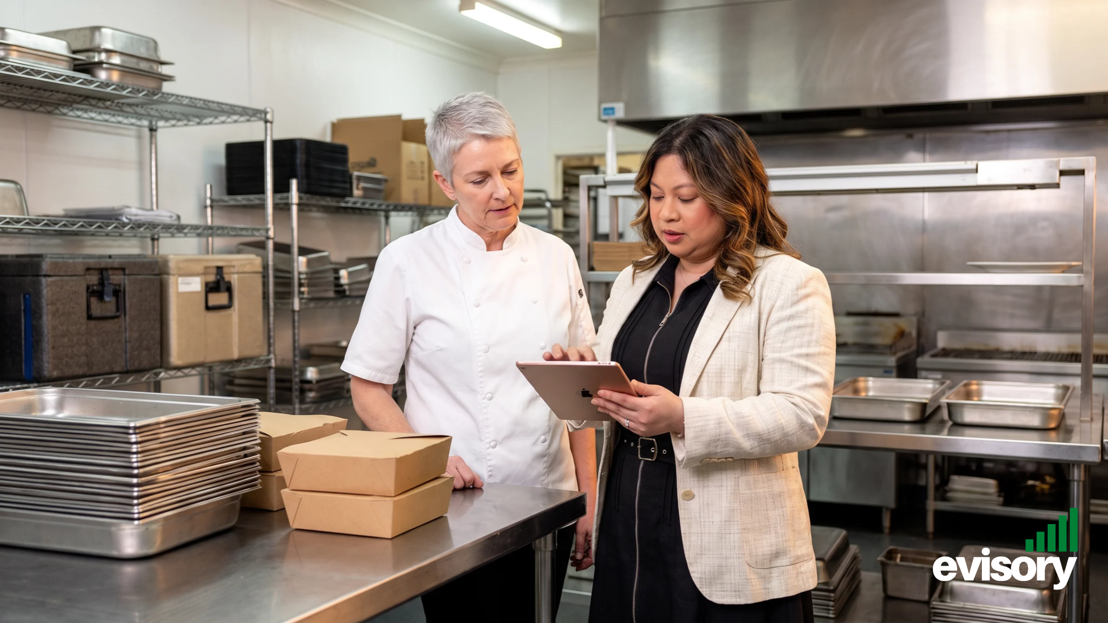 Linda Dang reviewing financial reports on a tablet alongside a head chef standing at a stainless steel prep counter in a commercial kitchen.