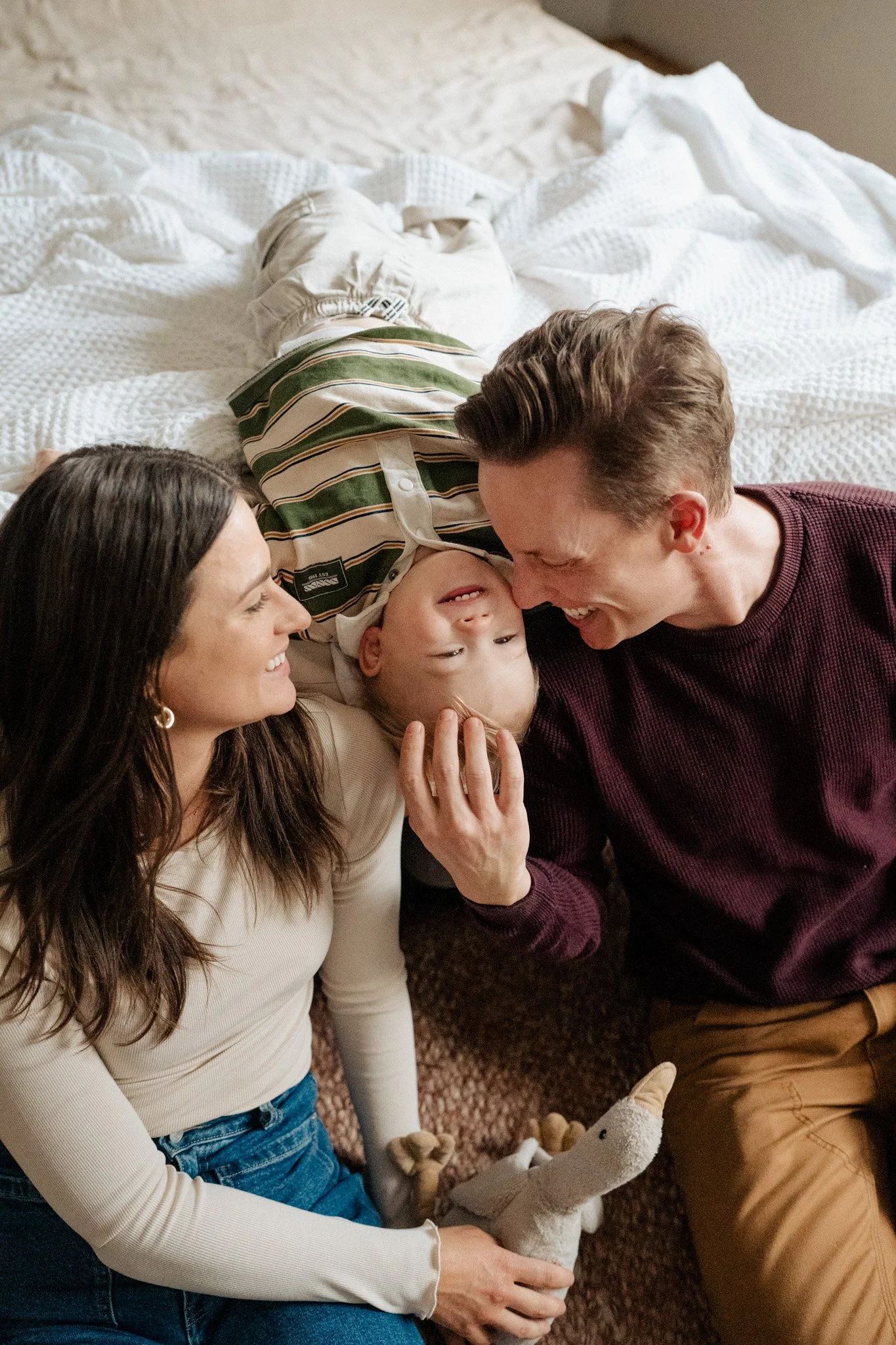 Parents smiling and kissing their laughing baby while lying together on a bed with a stuffed animal during an in-home family photography session in Melbourne.
