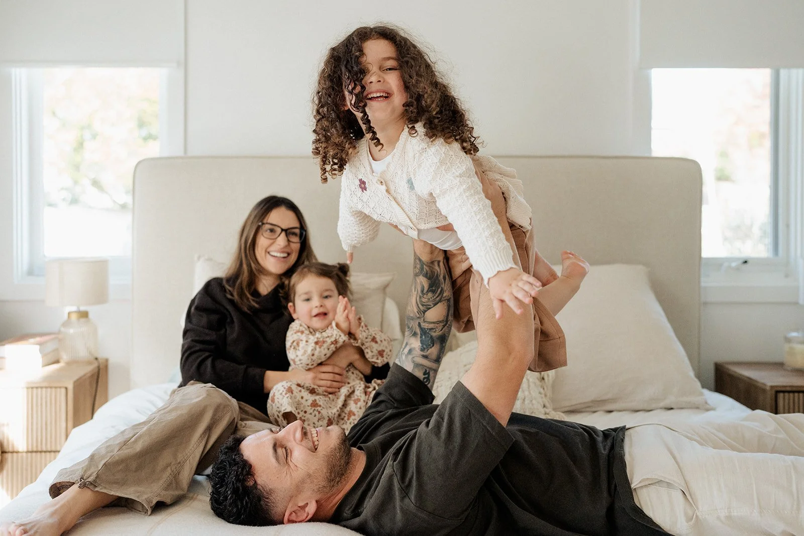 A girl being picked up by her dad and "flying" in the air on the parents bed. Mum and sister look on. all wth smiles during an in-home family photography session in melbourne