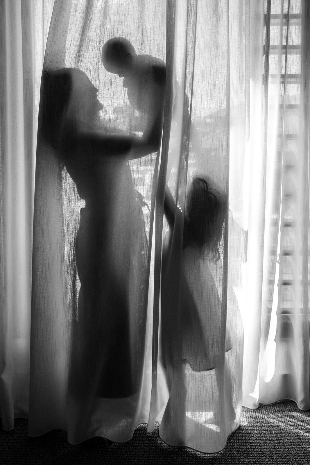 Black-and-white silhouette of a mother holding a baby behind sheer curtains, with a child standing beside them during an in-home motherhood photography session in Melbourne.