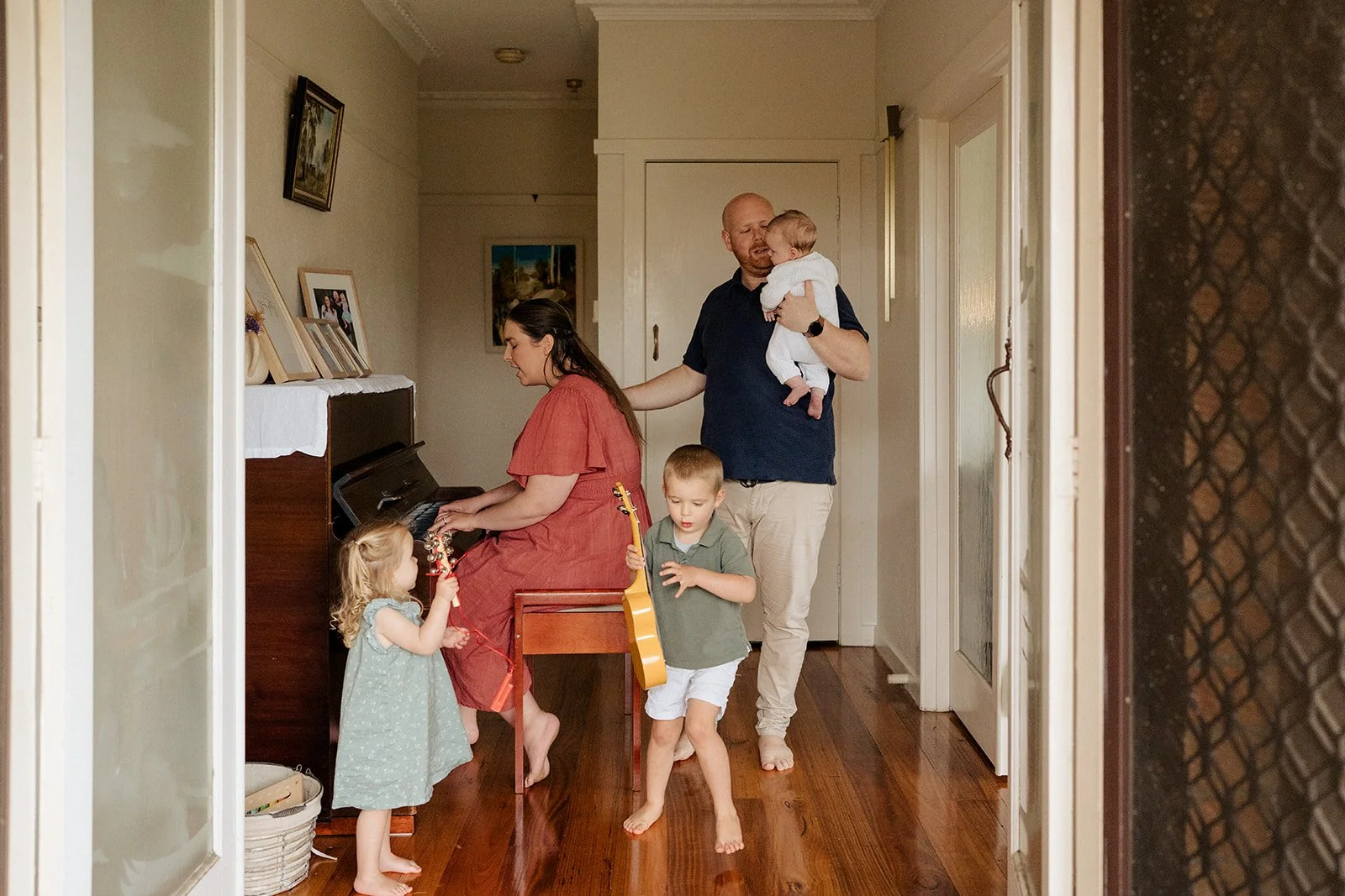 A mom sitting at the piano playing songs, while her husband hold her 1 month old baby boy. An older boy dances around holding a guitar and a two year old girl shakes a musical instrument. At a in-home newborn photography session in Melbourne