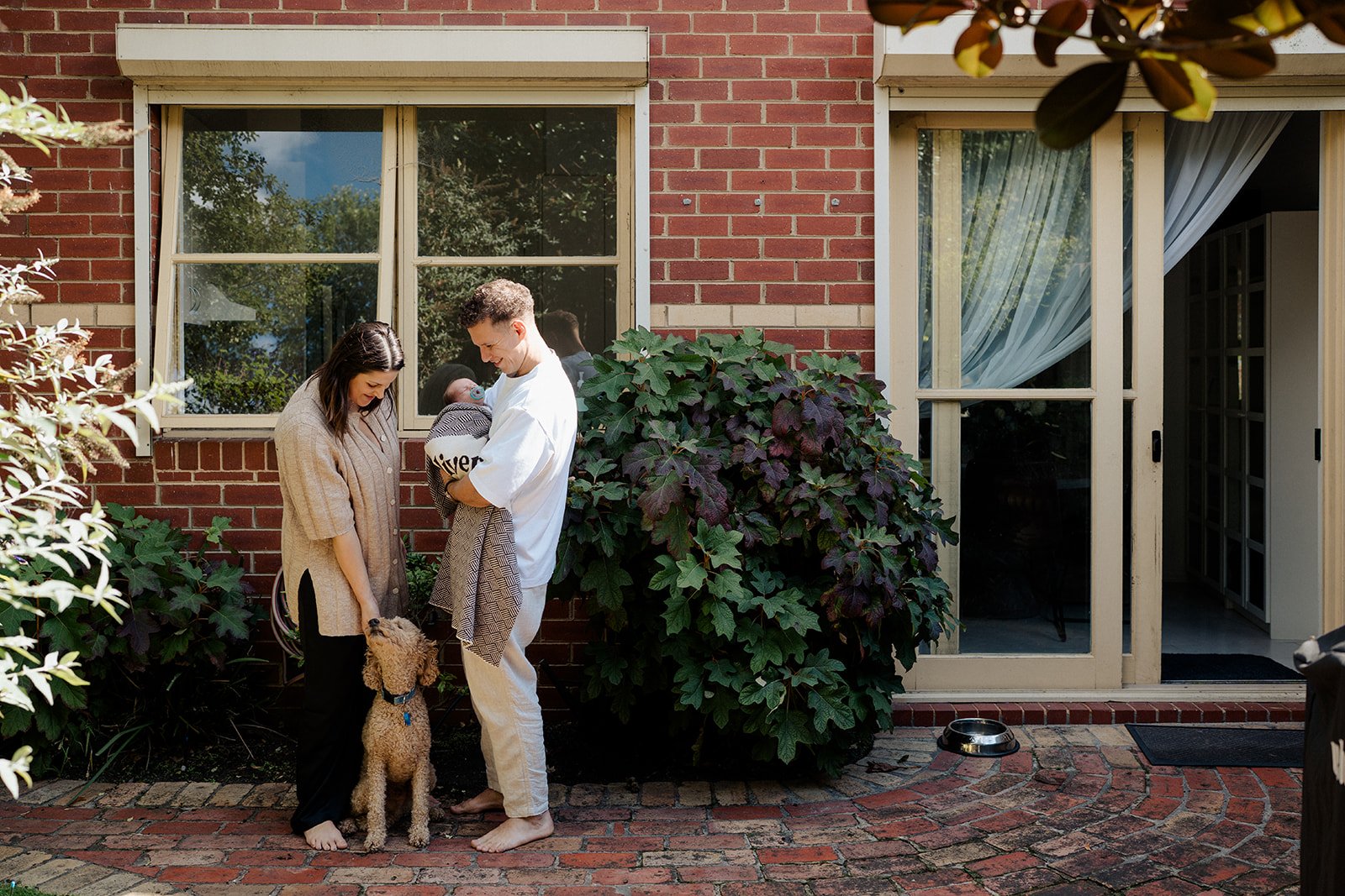 A newborn baby with mum, dad and dog standing outside their home in their backyard during a in-home newborn session in Melbourne