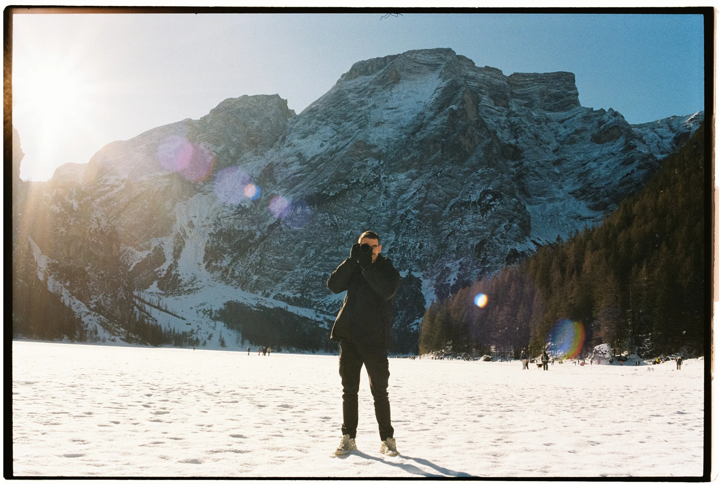 Persona tomándose una foto con fondo de montañas nevadas y nieve en el suelo, con otros visitantes en la distancia.