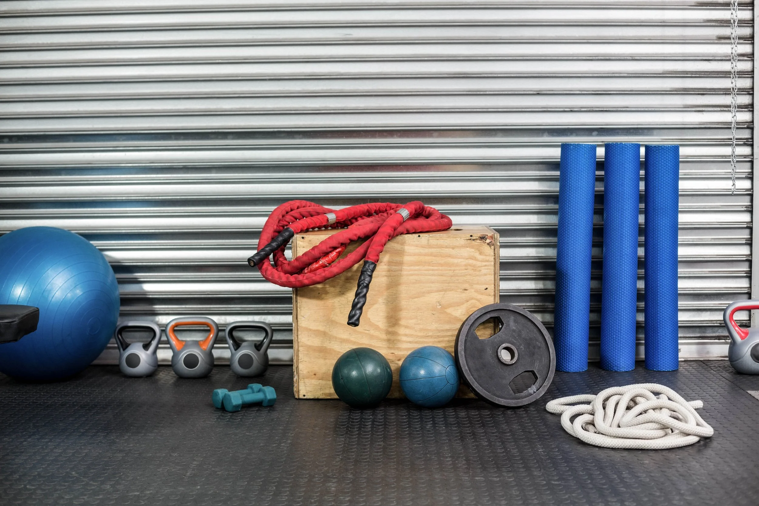 Gym equipment including a blue stability ball, kettlebells, resistance bands, foam rollers, weight plates, a battle rope, dumbbells, and battling ropes, all arranged against a silver metal wall.