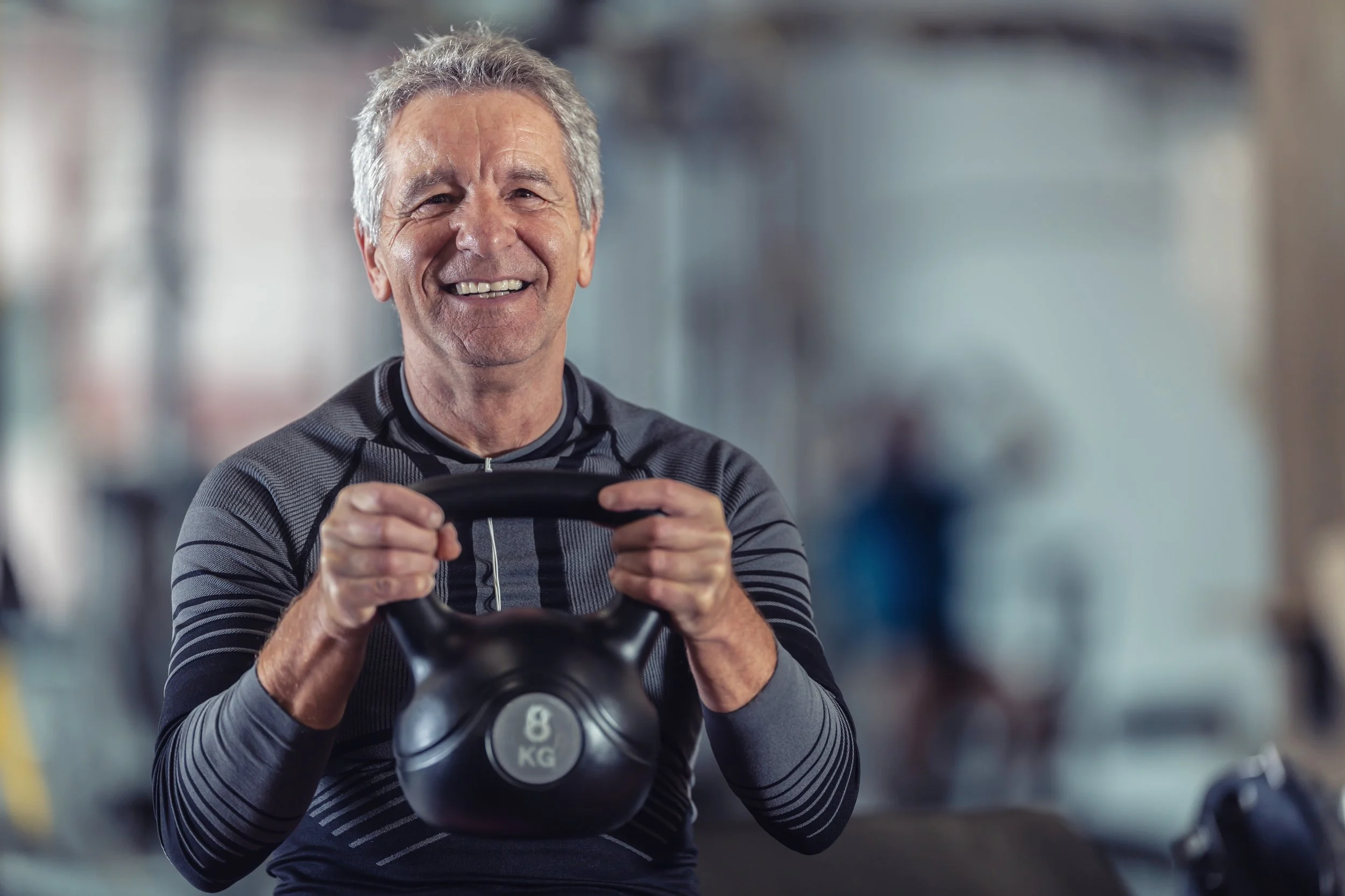 Older man smiling while holding a kettlebell in a gym.