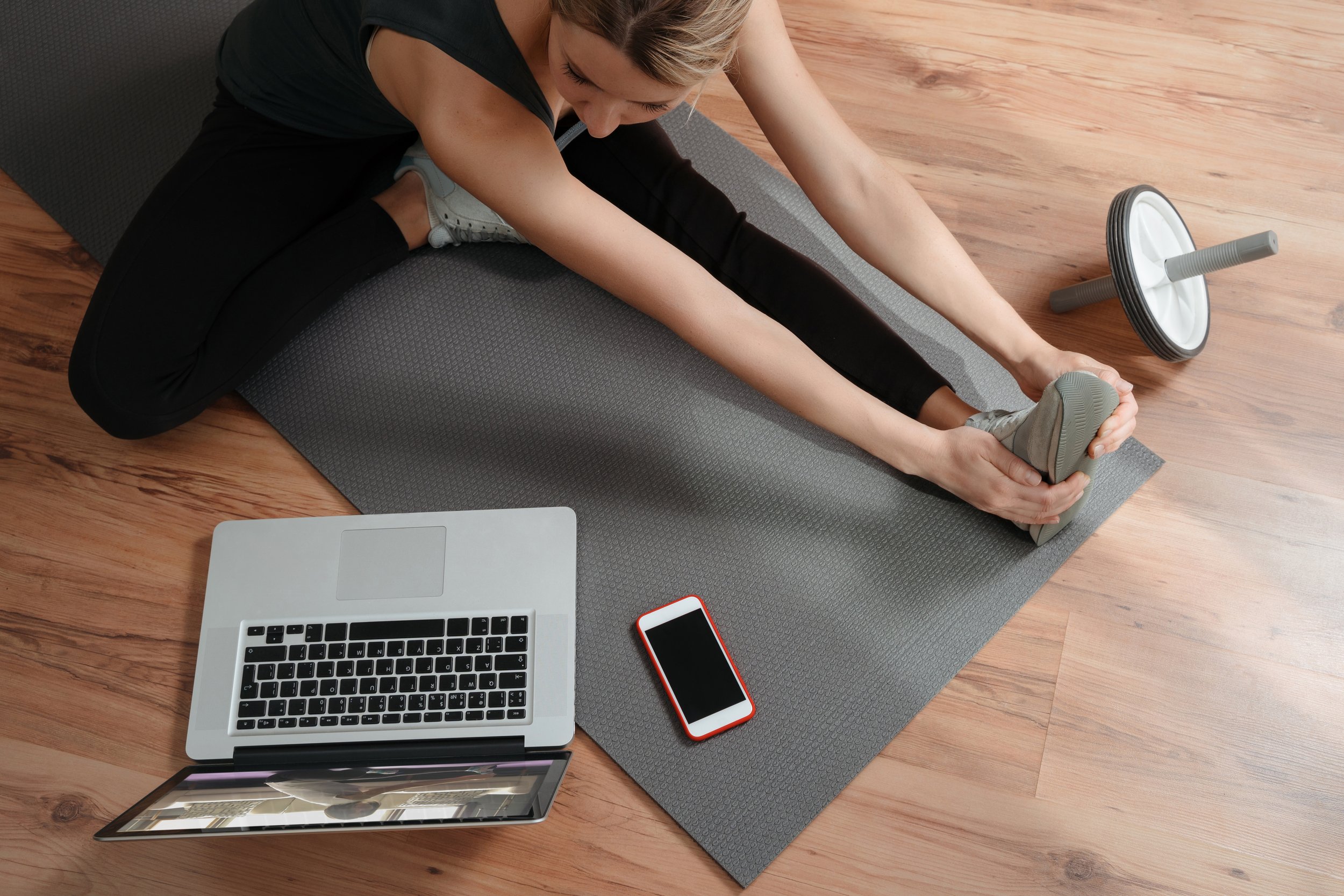 A woman in black workout clothes doing a seated forward stretch on a yoga mat, with a foam roller nearby, on a hardwood floor, surrounded by a laptop and a smartphone.