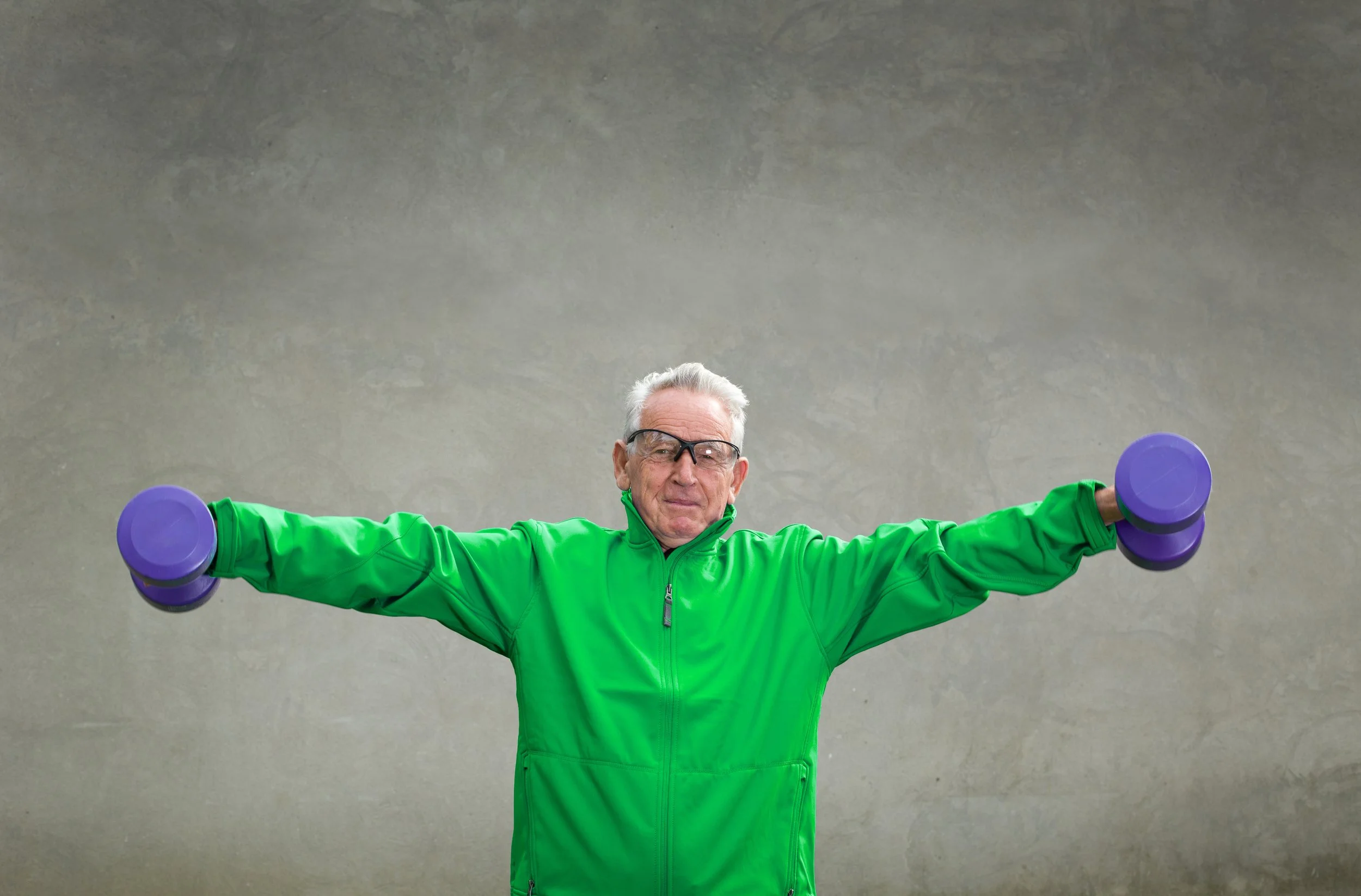 An elderly man wearing glasses and a bright green athletic jacket is lying on his back on a concrete floor. He is holding two purple dumbbells with extended arms, performing a workout.