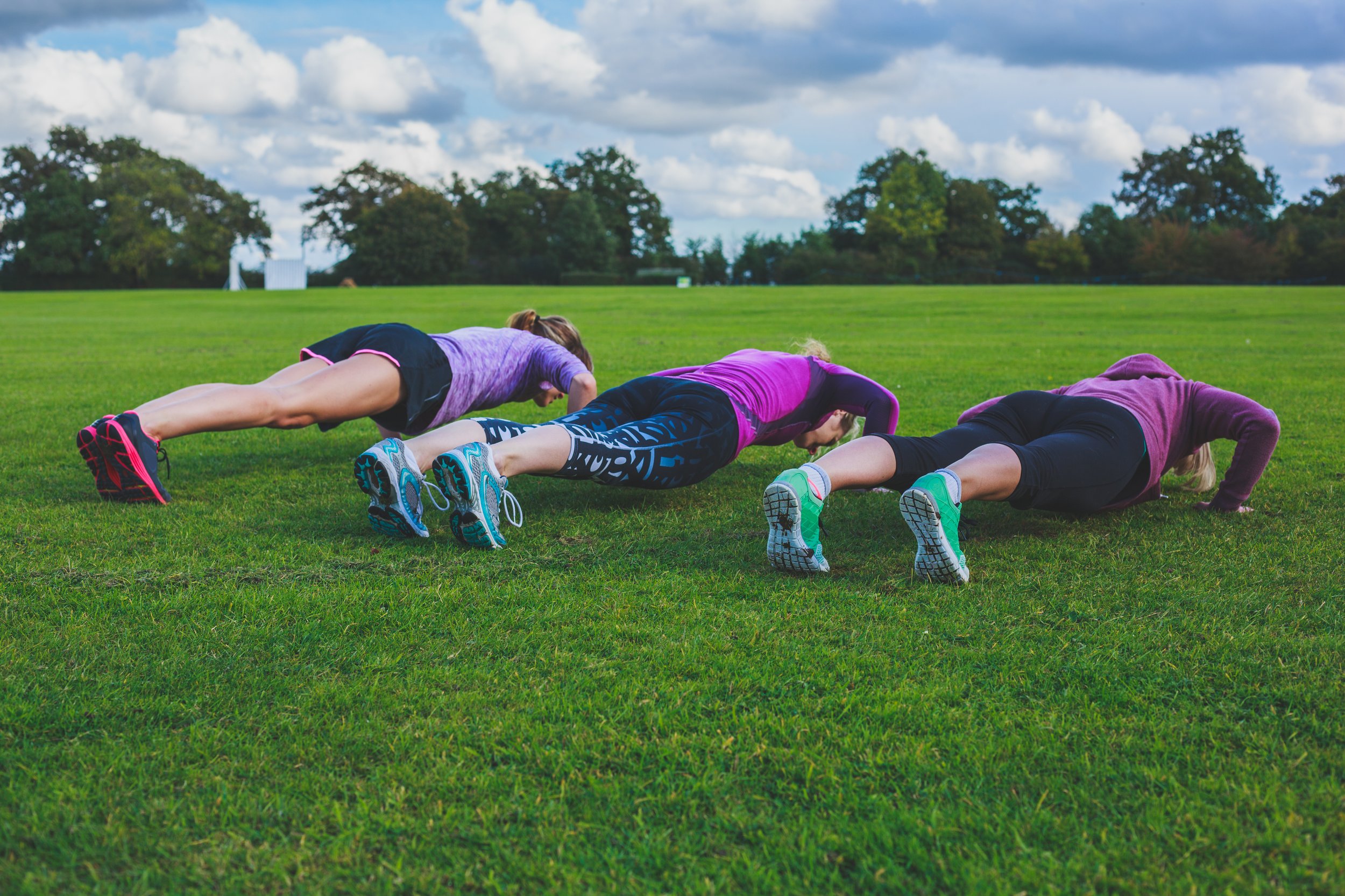 Three girls doing push-ups on a grassy field with trees and a cloudy sky in the background.
