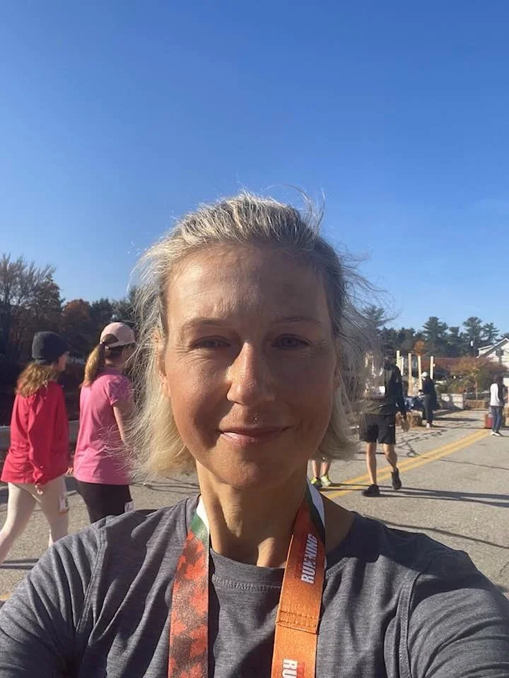 A woman taking a selfie outdoors during a race or event, with other participants in colorful athletic clothing in the background, under a clear blue sky.