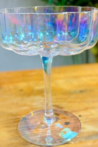 A clear glass cocktail or dessert glass with an iridescent finish, placed on a wooden surface with a blurred green background.