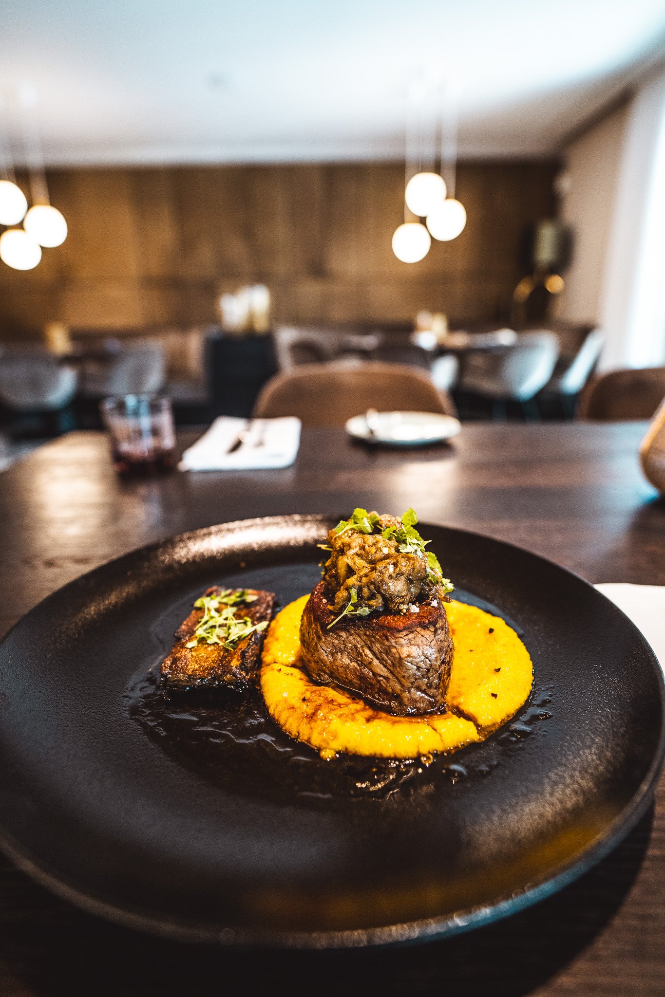 Auf einem schwarzen Teller liegt ein Steak mit einer Kräuterkruste, serviert auf einem cremigen Püree. Im Hintergrund ein gehobener Restaurantraum.