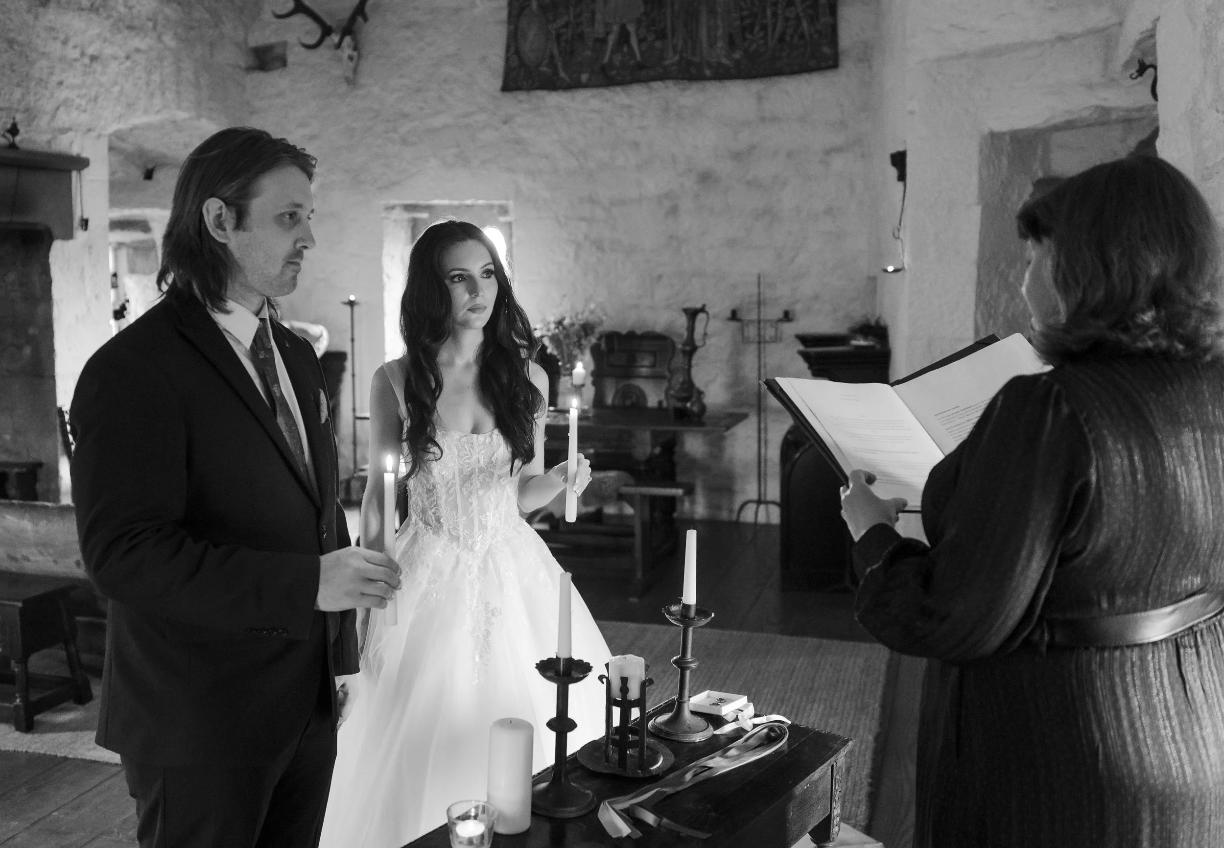 A black and white photo of a wedding ceremony inside a rustic stone building. A man in a suit and a woman in a wedding dress hold candles, standing side by side, facing a woman holding a book, who appears to be officiating the ceremony.