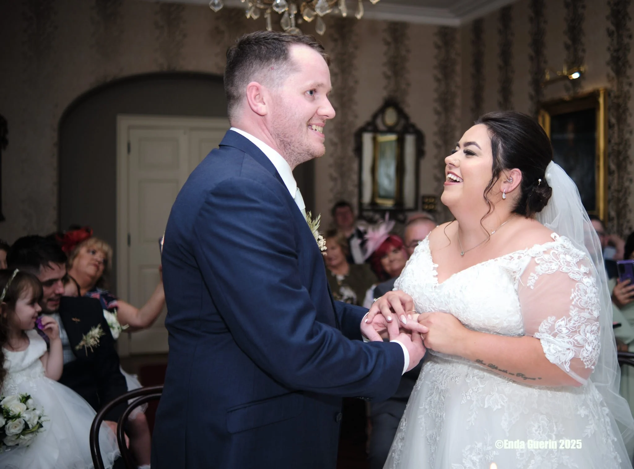 A bride and groom holding hands during their wedding ceremony, smiling at each other in an elegant indoor setting with wedding guests in the background.