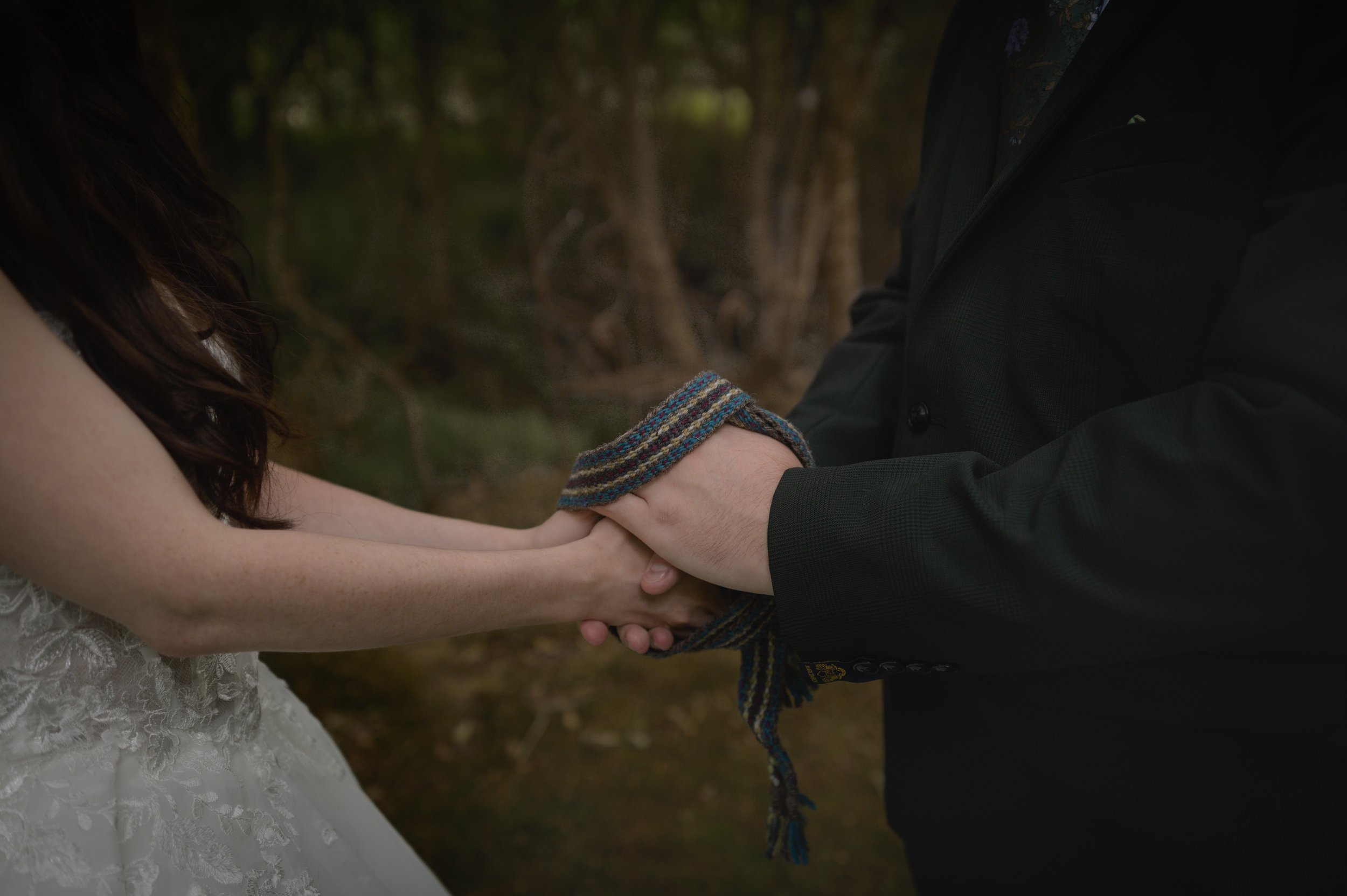 A man and a woman holding hands outdoors, the woman is wearing a white lace dress, and the man is wearing a dark jacket. Their hands are clasped together, and the woman's wrist is adorned with a colorful woven bracelet.
