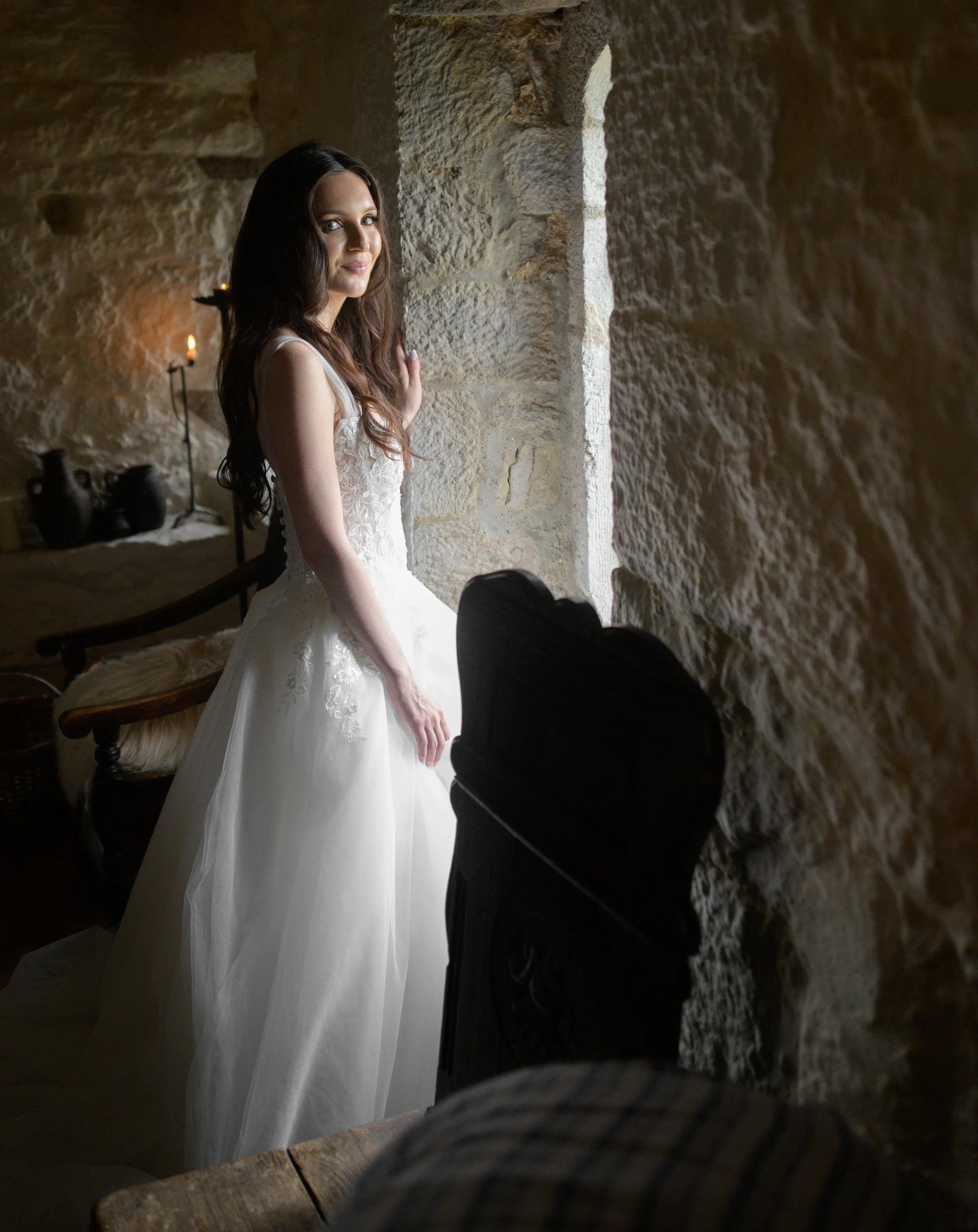 A young woman in a white dress looking out a window in a stone-walled room with candles in the background.
