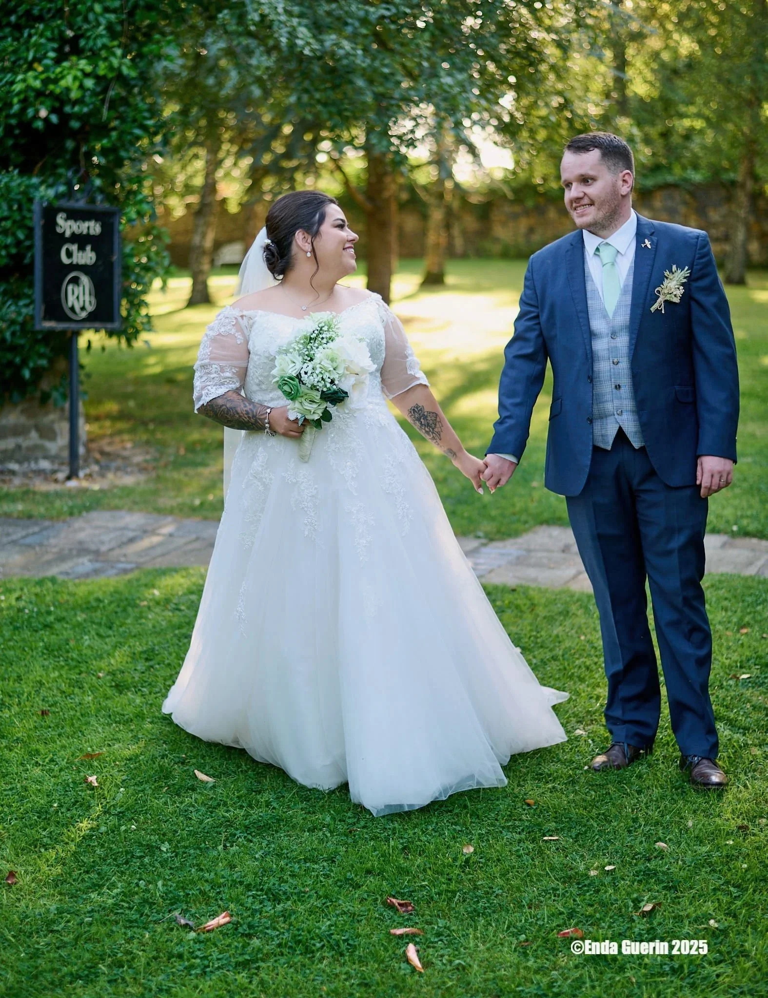 A bride and groom holding hands, standing on a grassy area outdoors with trees in the background, smiling at each other during their wedding.