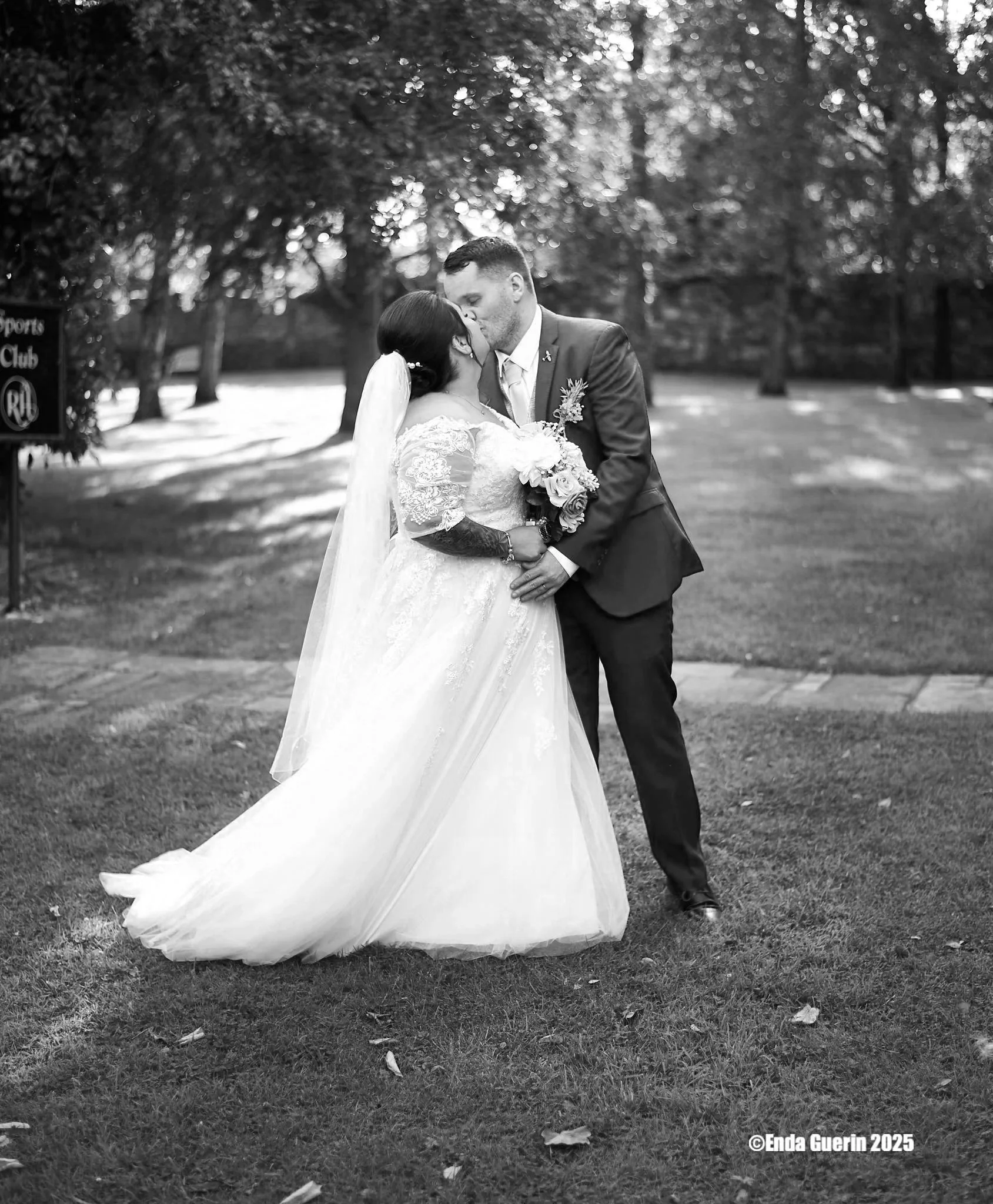 A bride and groom kiss outdoors on a grassy area, surrounded by trees, during a wedding. The bride wears a white lace wedding dress, and the groom wears a navy blue suit. The bride holds a bouquet of white and green flowers, and a veil flows behind h