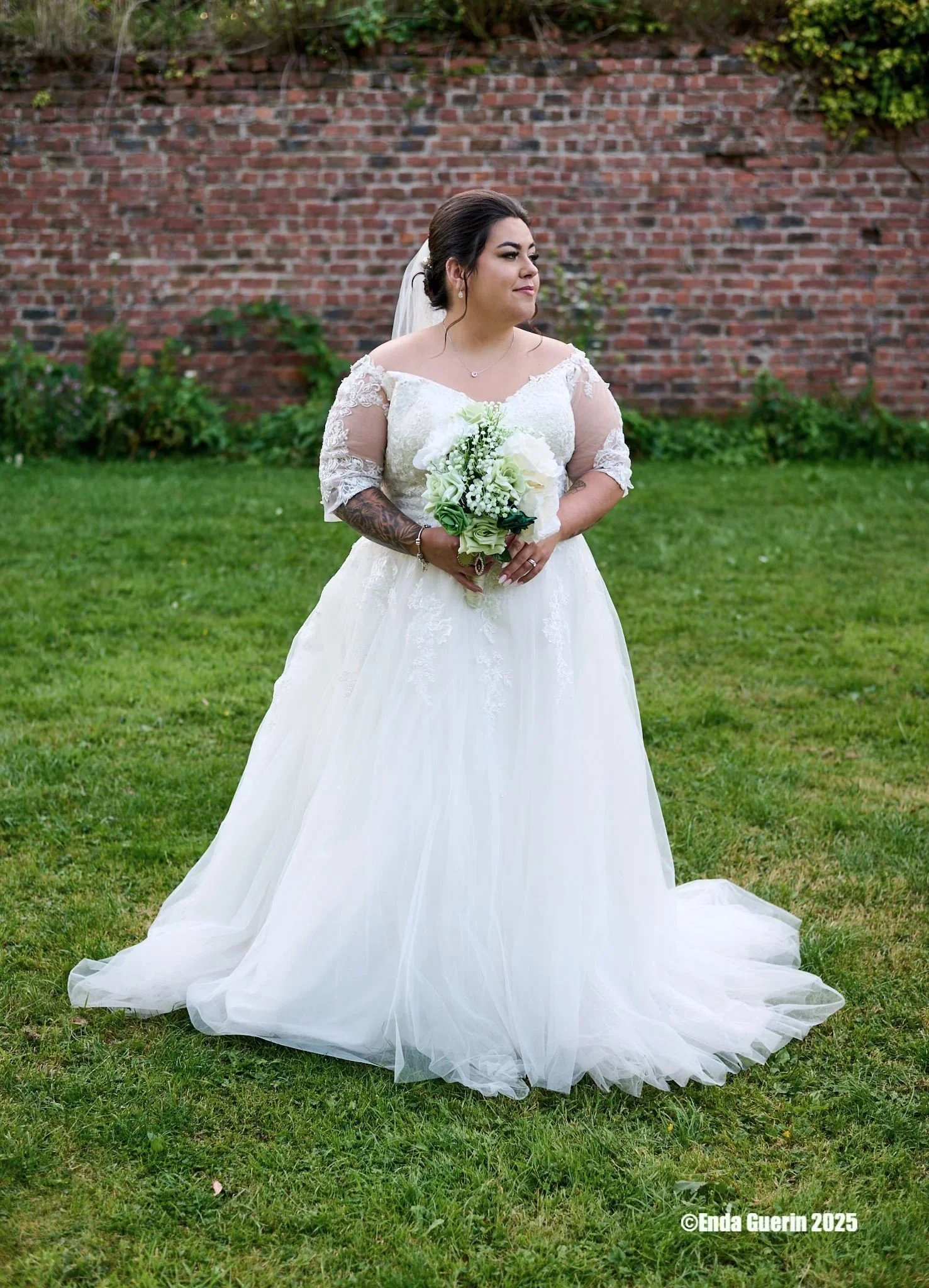 A bride standing on a grassy lawn holding a bouquet of white and green flowers, wearing a white lace wedding dress with sheer lace sleeves, dark hair styled up, and a veil, with a brick wall and greenery in the background.