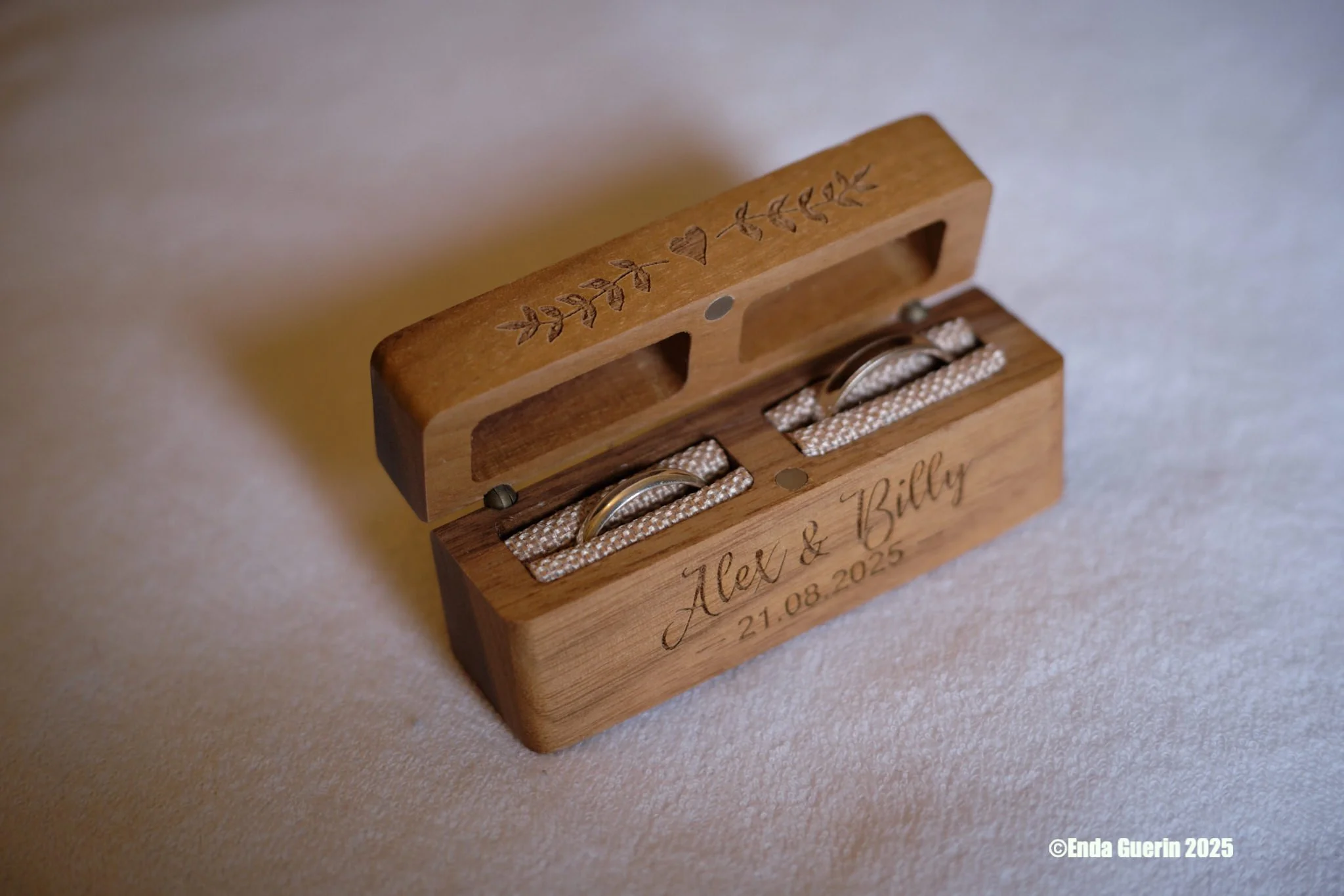 Wooden ring box with engraved decorations and the names 'Alex & Billy' along with the date '21.08.2025'; contains two wedding rings on beige fabric cushions.