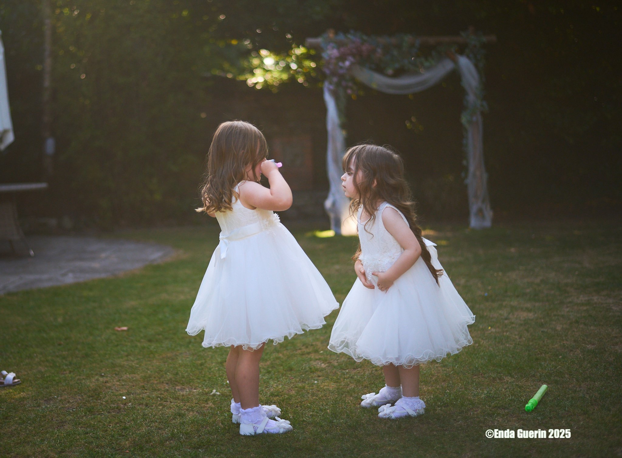 Two young girls in white dresses face each other on a grassy lawn, with a decorated arch behind them, during what appears to be a wedding or special event, under warm light.