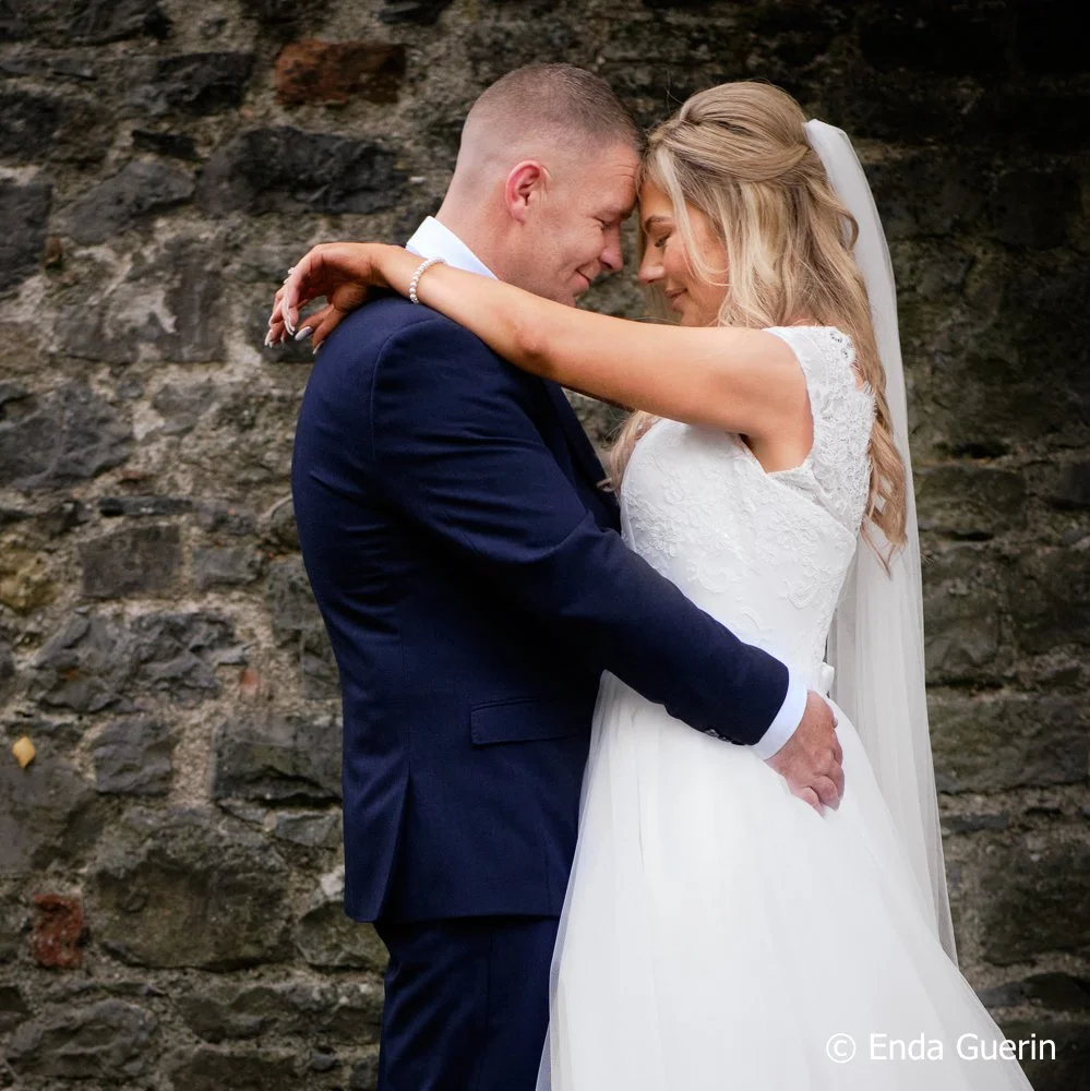 A bride and groom embrace tenderly with foreheads touching, standing against a stone wall, during their wedding.