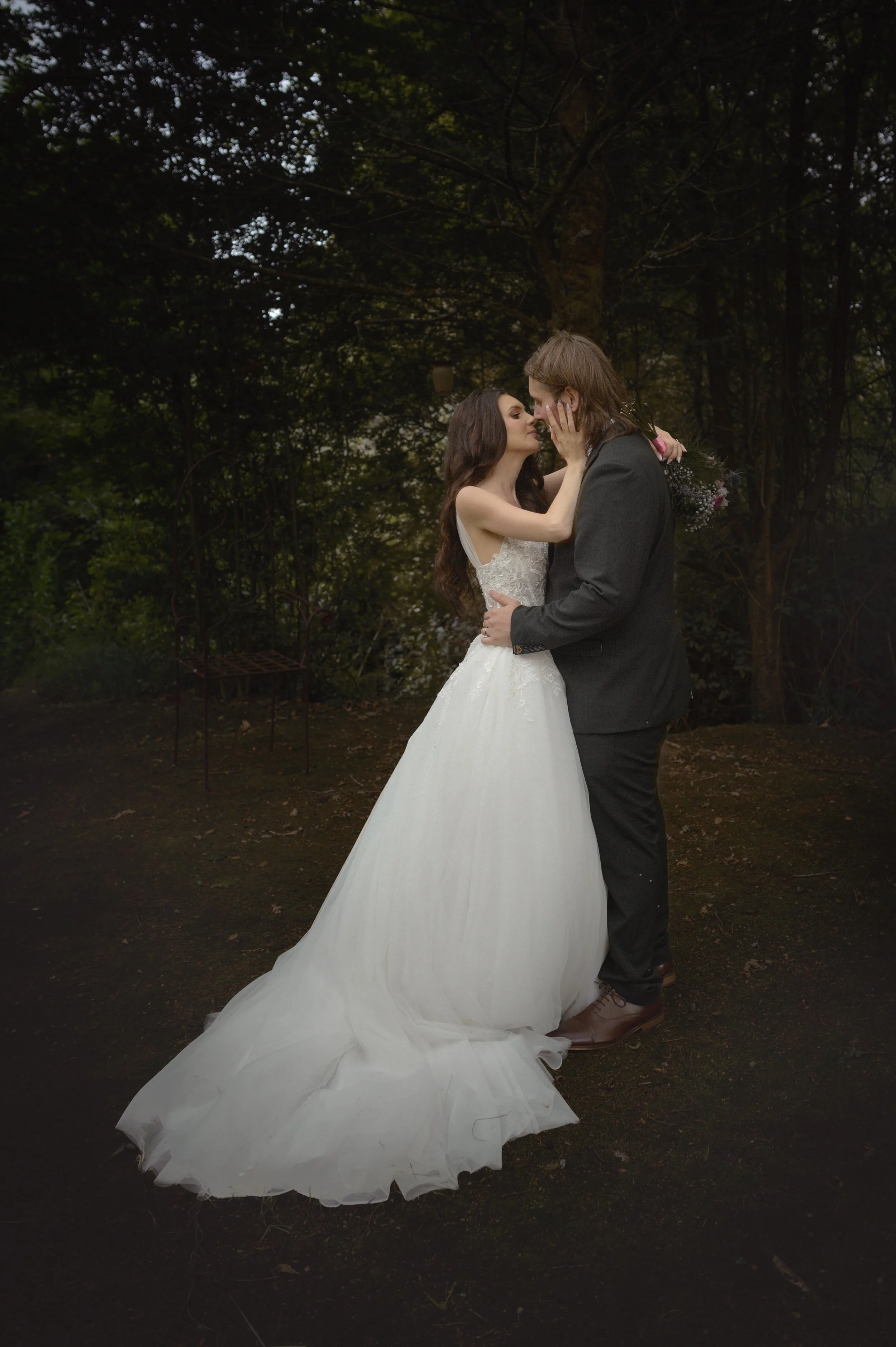 A bride and groom standing close together outdoors, embracing and about to kiss, with the bride holding a bouquet of flowers, in a wooded area during daytime.