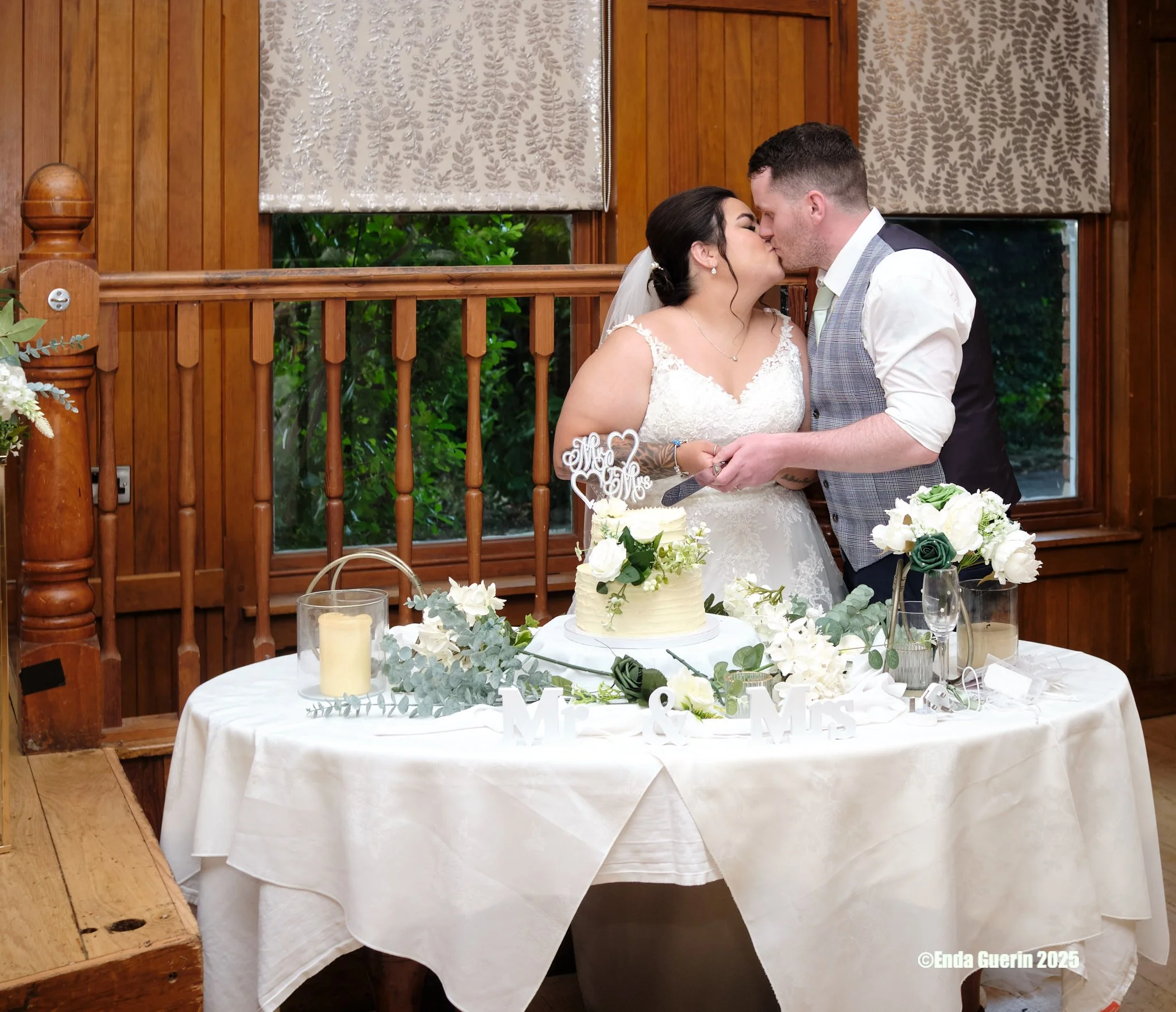 Bride and groom kissing at their wedding reception, with a table of decorations and a wedding cake in front of them.