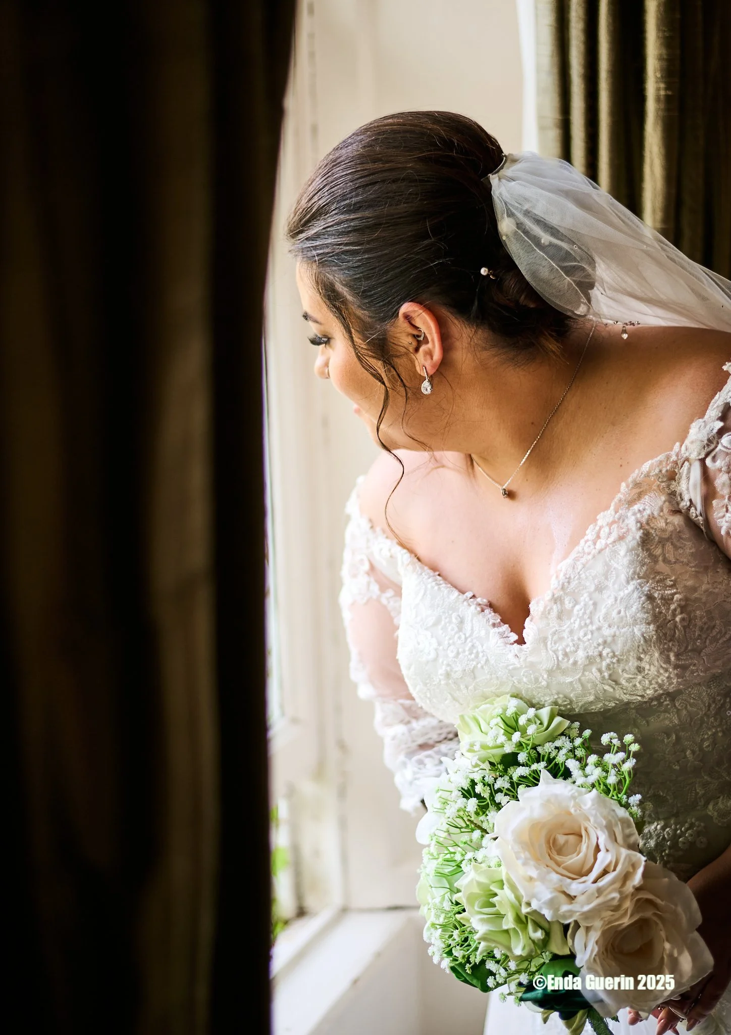 A bride in a white lace wedding dress looking out a window while holding a bouquet of white roses and greenery.