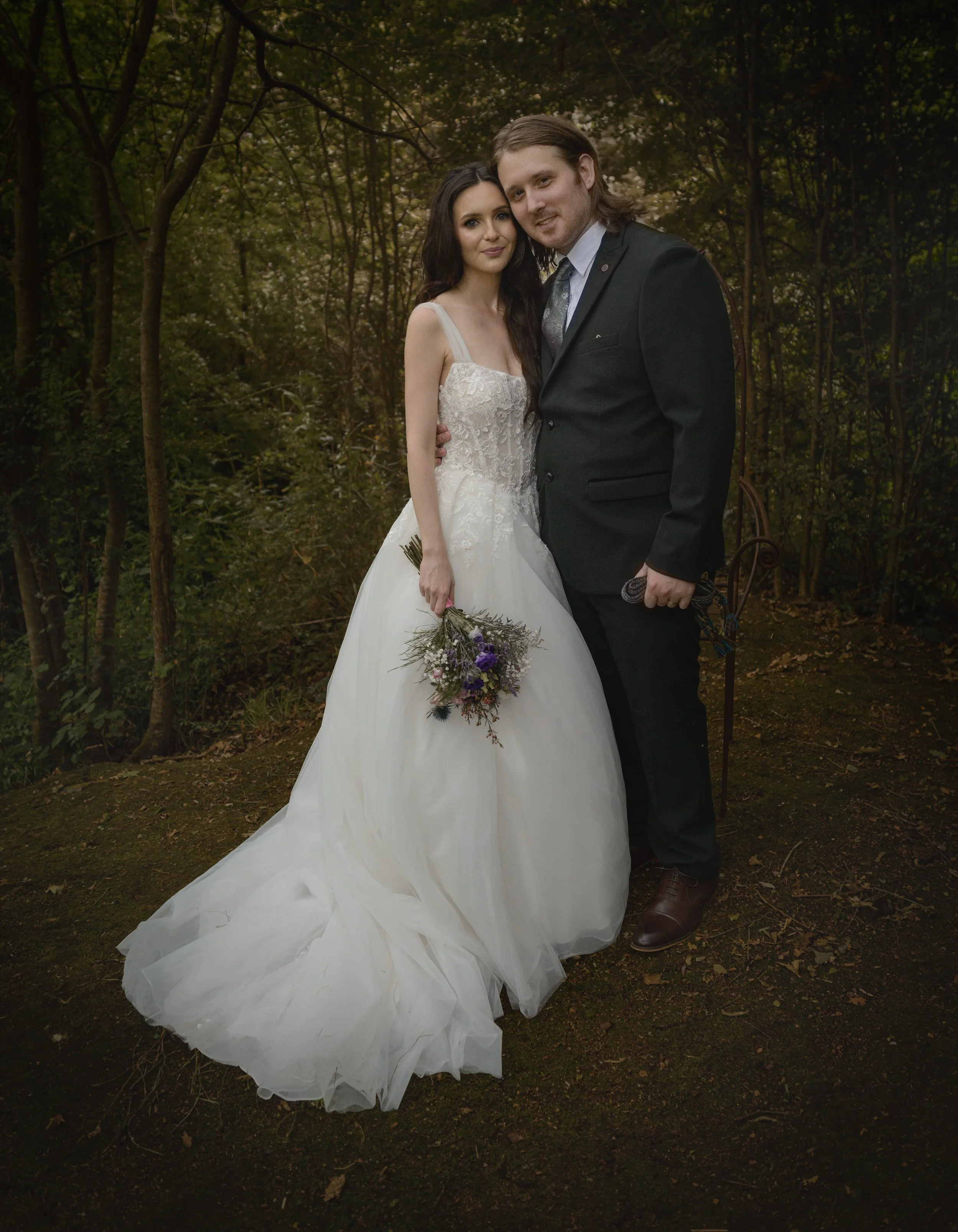 A bride and groom posing together outdoors in a wooded area, with the bride holding a bouquet of flowers. The bride wears a white wedding dress and the groom a dark suit. They are close together, looking at the camera.