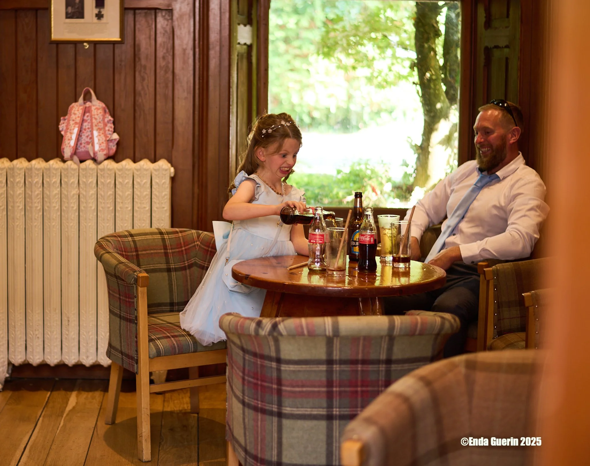 A young girl in a white dress is pouring a drink for a man sitting at a table with various bottles and glasses in a cozy wooden room with outdoor greenery visible through the window.