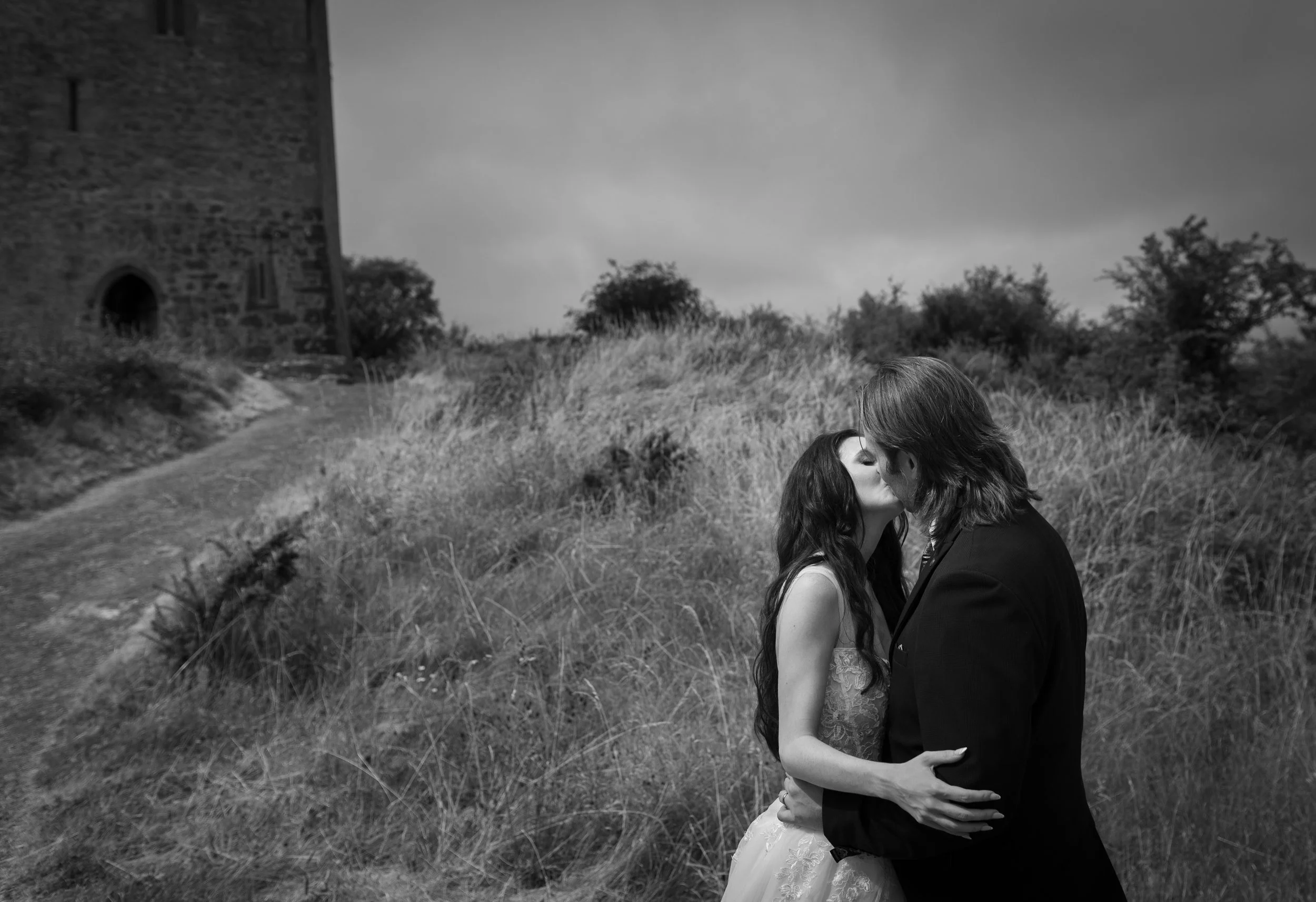 A black and white photograph of a couple in a passionate embrace, standing on a grassy area with a historic stone building in the background. The woman has long, dark hair and is wearing a dress, while the man has medium-length hair and is wearing a 