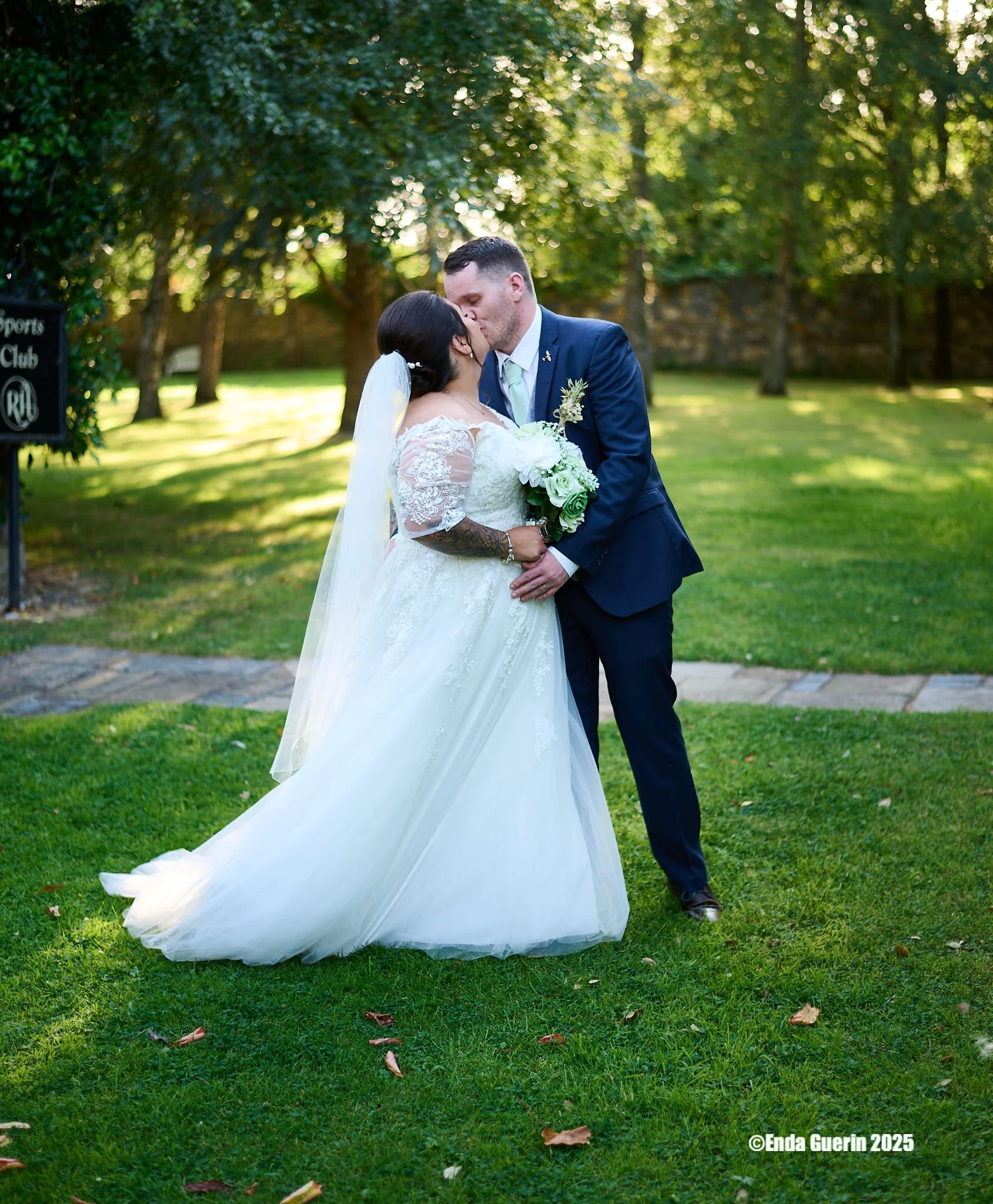 A bride and groom kiss outdoors on a grassy area, surrounded by trees, during a wedding. The bride wears a white lace wedding dress, and the groom wears a navy blue suit. The bride holds a bouquet of white and green flowers, and a veil flows behind h