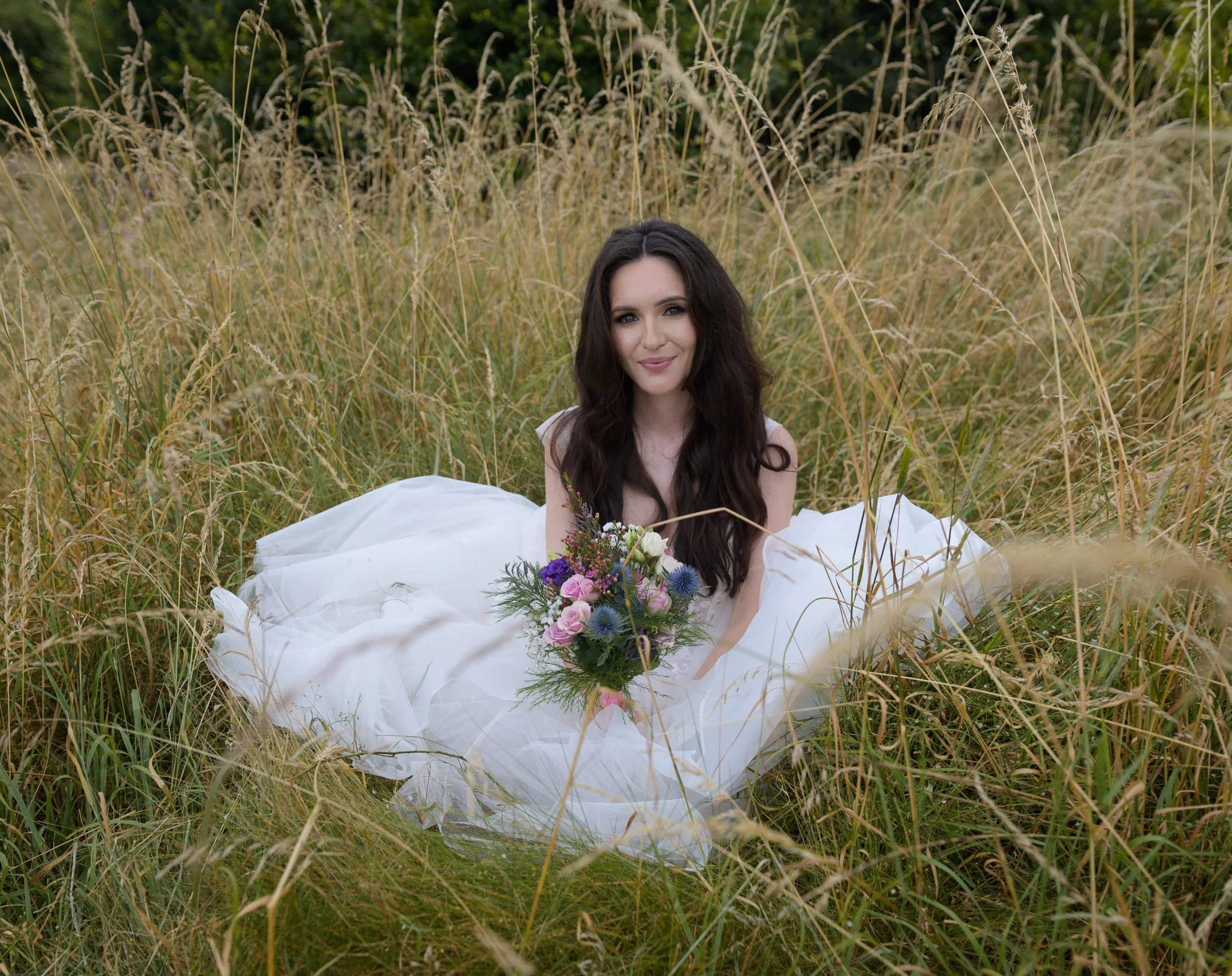 A woman with long dark hair, smiling, sitting in a field of tall grass, holding a bouquet of flowers on a white fabric.