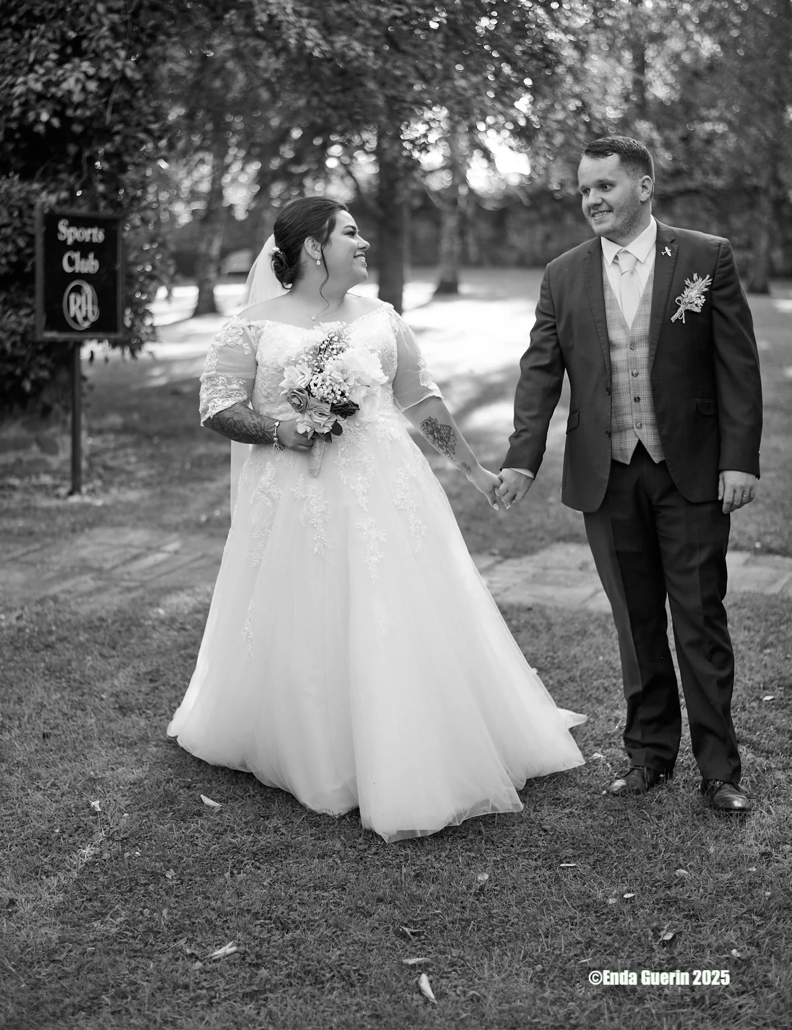 A bride and groom holding hands and smiling at each other outdoors on a grassy area with trees in the background. The bride is wearing a lace wedding dress and holding a bouquet of flowers. The groom is dressed in a suit with a boutonniere. The photo