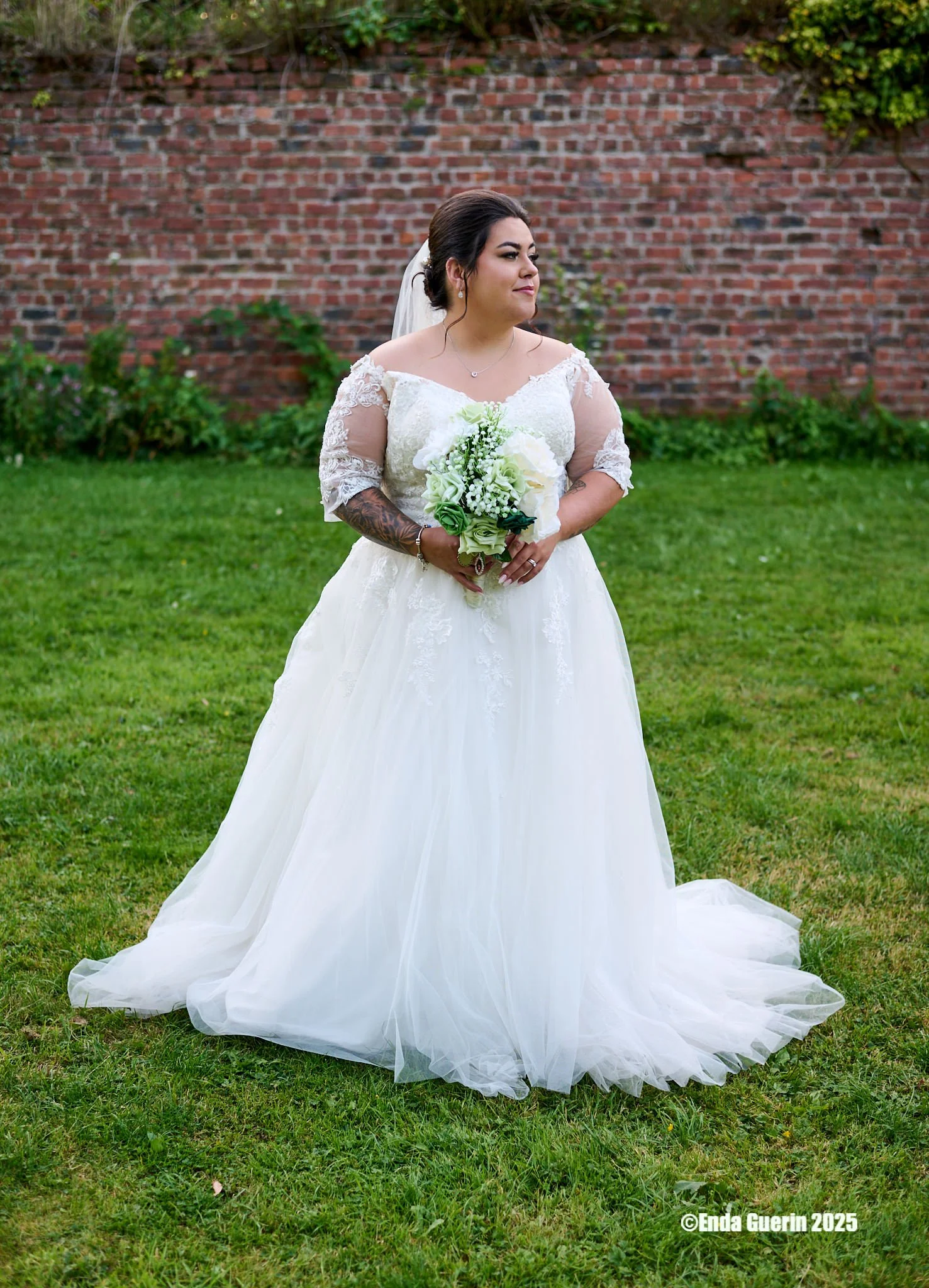 A bride standing on a grassy lawn holding a bouquet of white and green flowers, wearing a white lace wedding dress with sheer lace sleeves, dark hair styled up, and a veil, with a brick wall and greenery in the background.